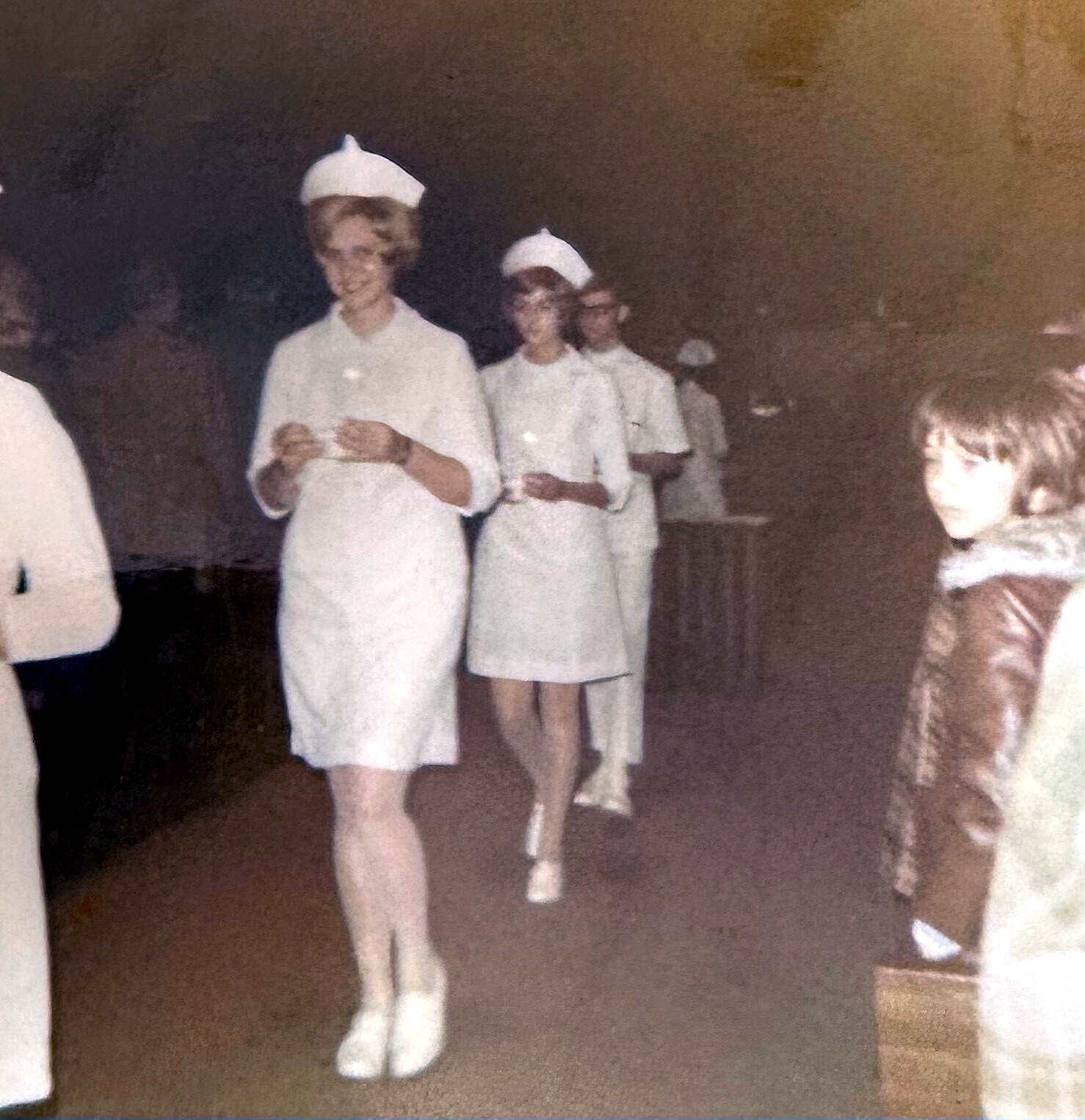 Vintage photo of young women in white dresses and nurse caps walking in a line indoors, likely a procession or ceremony.