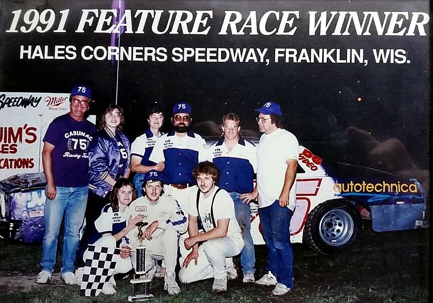 Group of racers and crew pose with trophy beside a race car at night, celebrating the 1991 feature race win at Hales Corners Speedway, Wisconsin