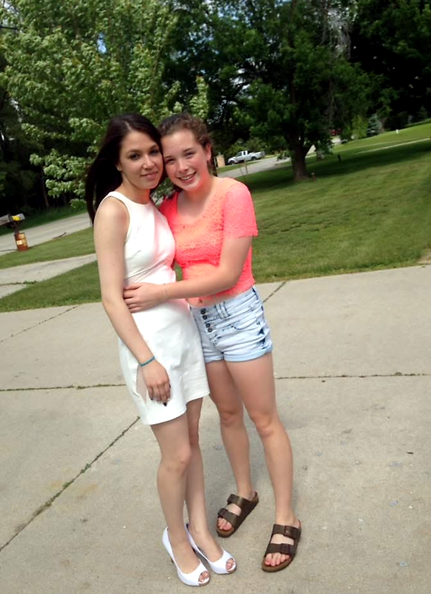 Two young women standing on a sidewalk in a park, arms around each other and smiling at the camera.