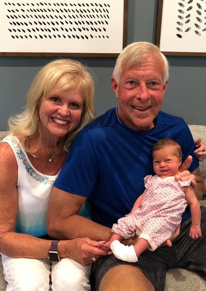 Smiling older couple sitting on a sofa, holding a newborn baby girl in pink pajamas in their arms.