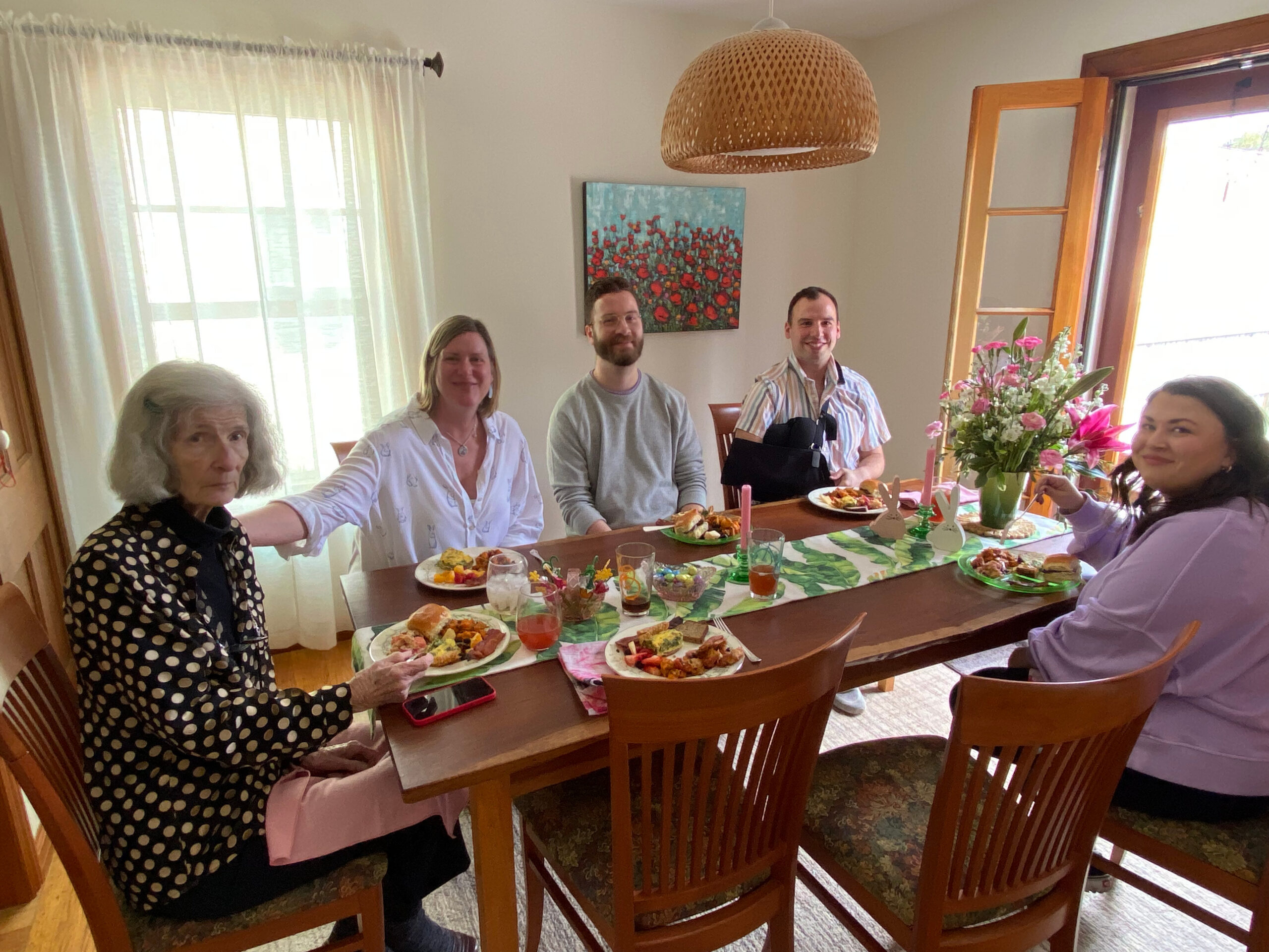 A group of six people seated around a dining table with snacks and drinks, smiling for the camera in a bright room.