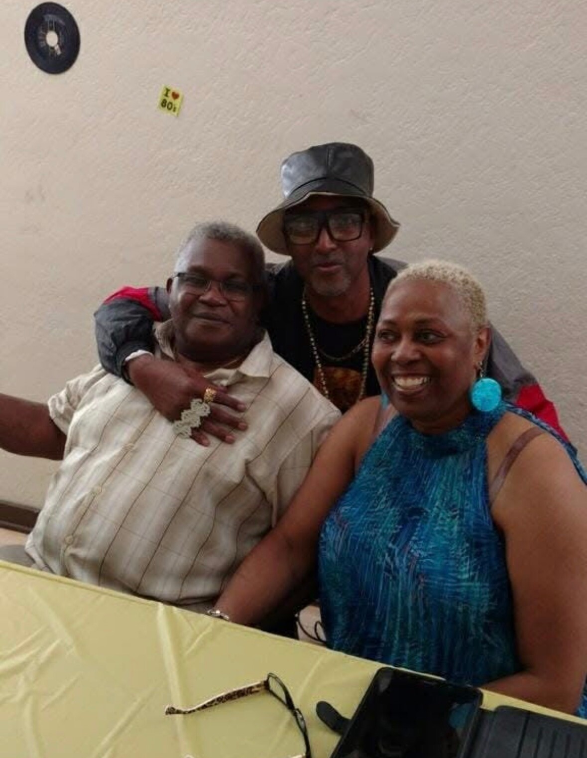 Three adults smile for the camera at a table; a man wearing a hat stands behind two seated companions with his arms around them, posing indoors.