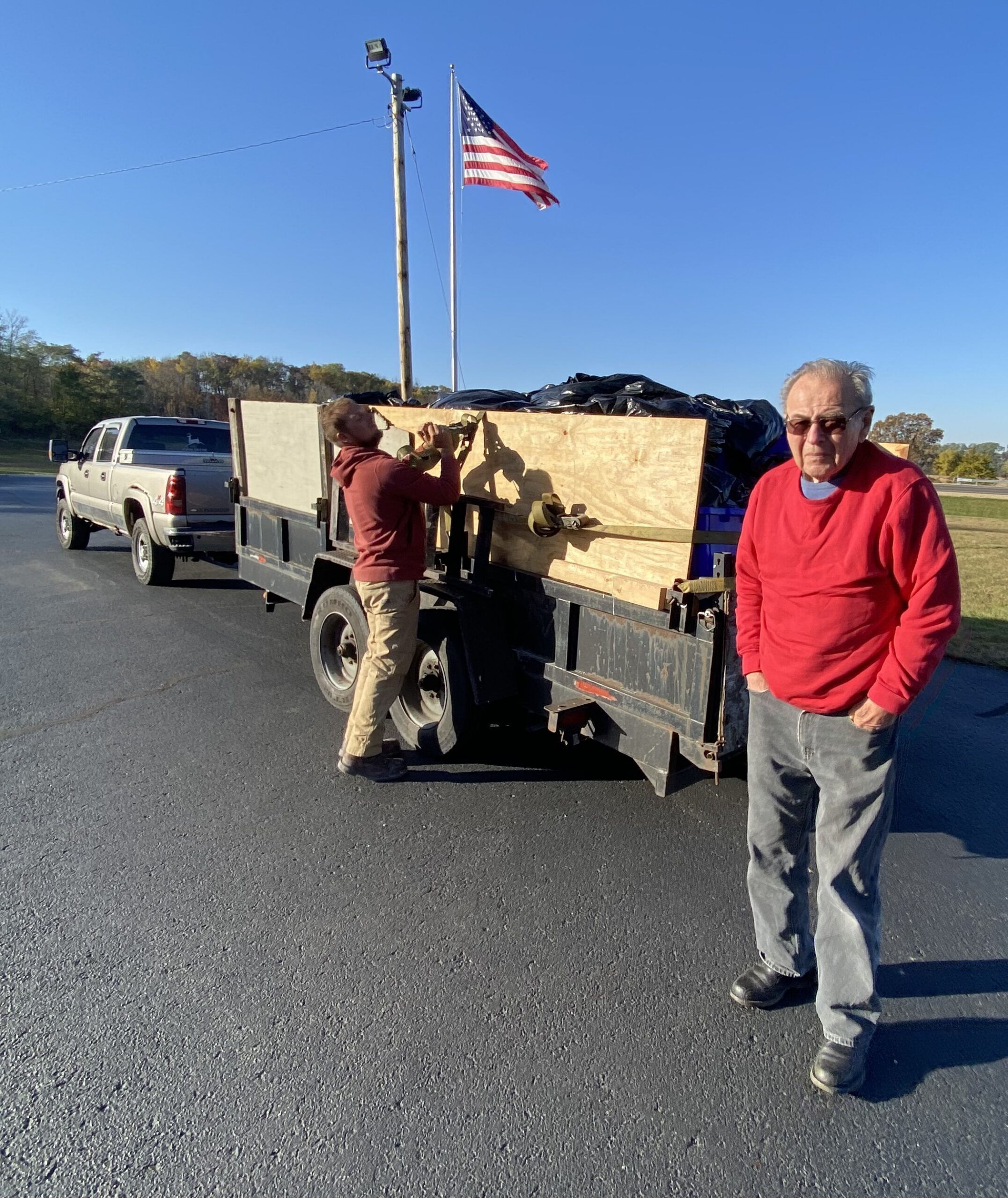 Two men standing near a flatbed trailer on a road, with an American flag flying on a pole nearby.
