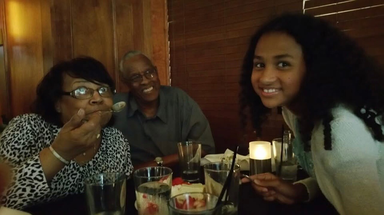 Three people sit at a restaurant table: an older woman with glasses tasting with a spoon, a smiling man beside her, and a smiling young woman on the right. A candle and drinks are on the table.