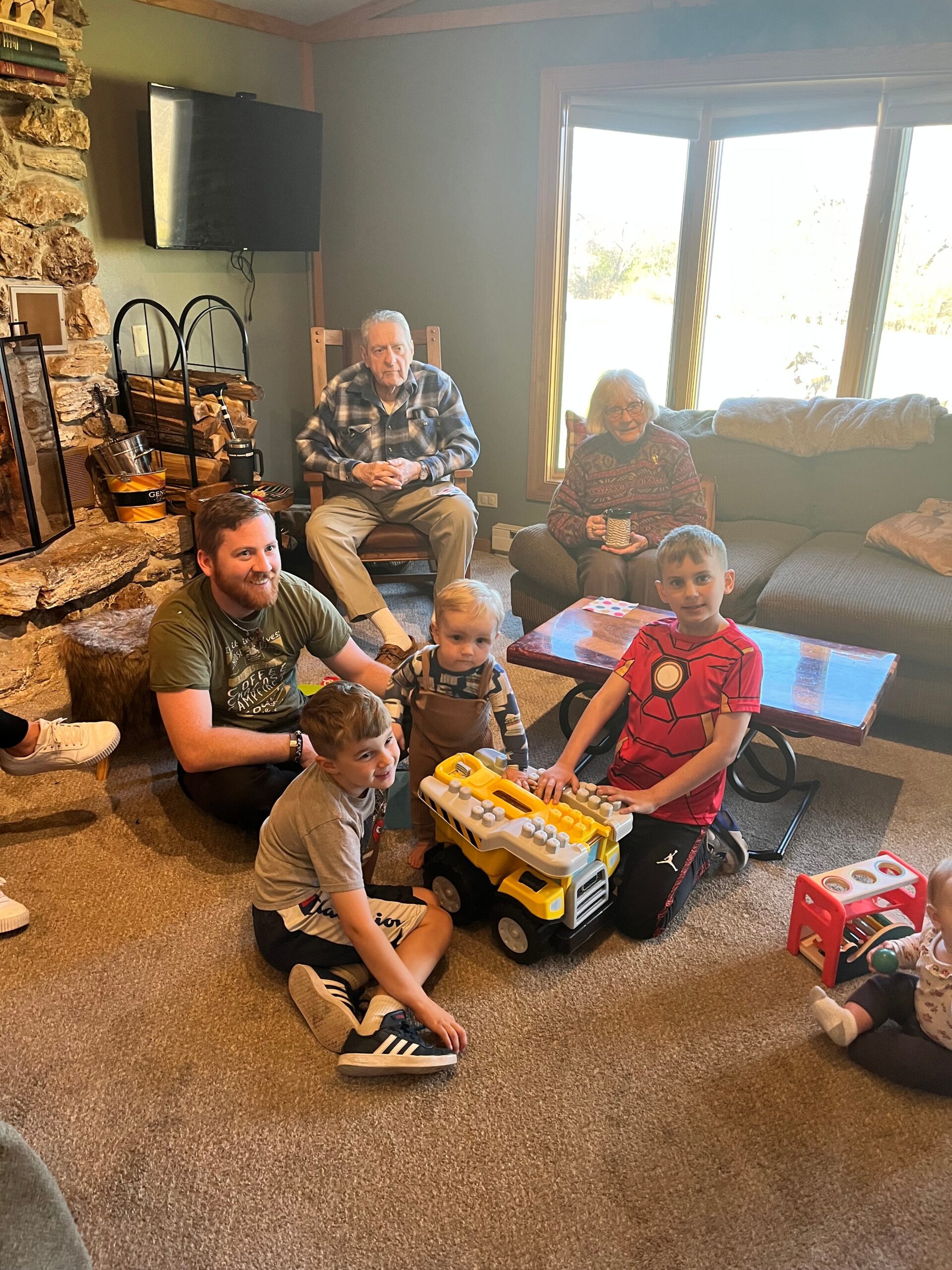Family group in a cozy living room: two seniors on the sofa while kids play with a large yellow toy truck nearby.