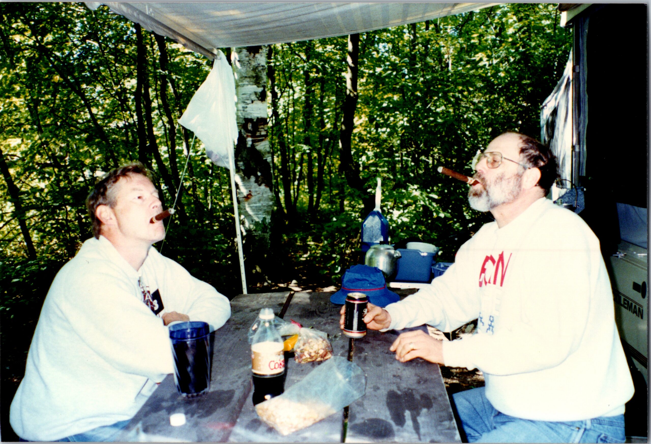Two men at a picnic table in a forested area, smoking cigars, with drinks and snacks on the table.