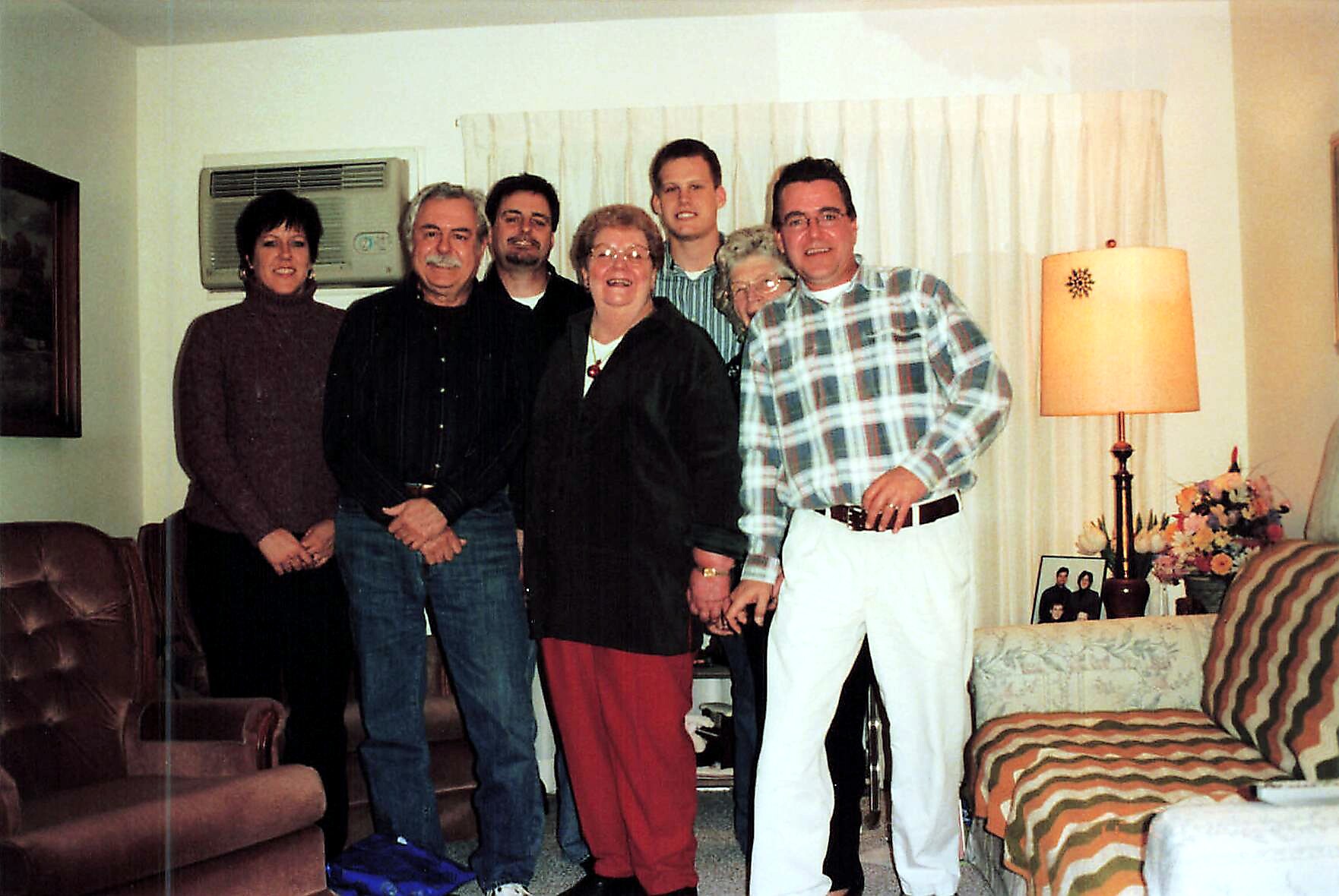 Seven adults pose together in a living room for a casual group photo, smiling at the camera.