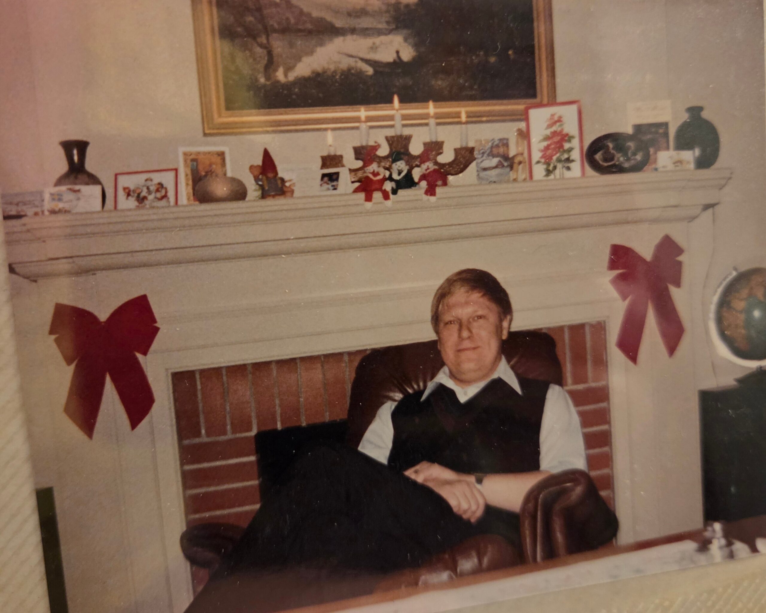 Older man sits on a chair in a living room, holiday decorations and cards on the mantel behind him.