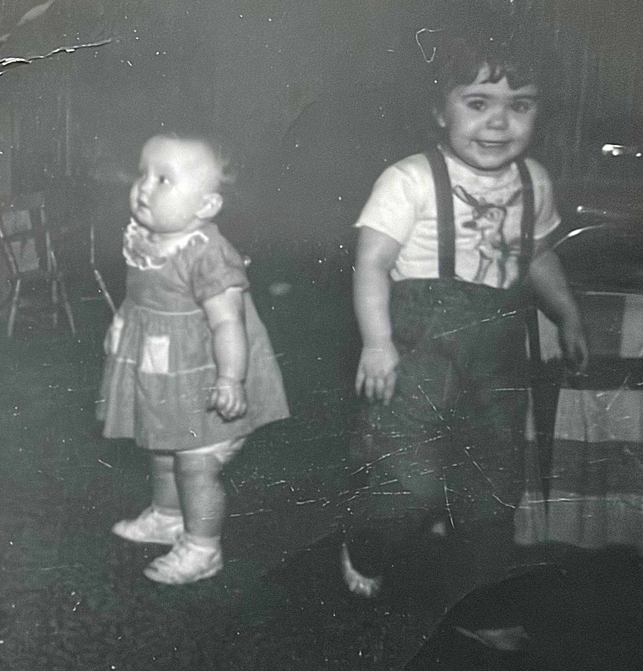 Two young children walk hand in hand outdoors in a grainy black-and-white photo; girl on left in a dress, boy on the right in suspenders and a striped shirt.