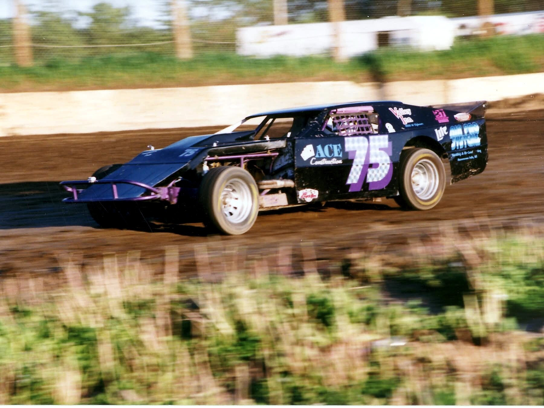Black and purple late-model stock car (number 75) speeding on a dirt oval, dirt flying off the rear tires.