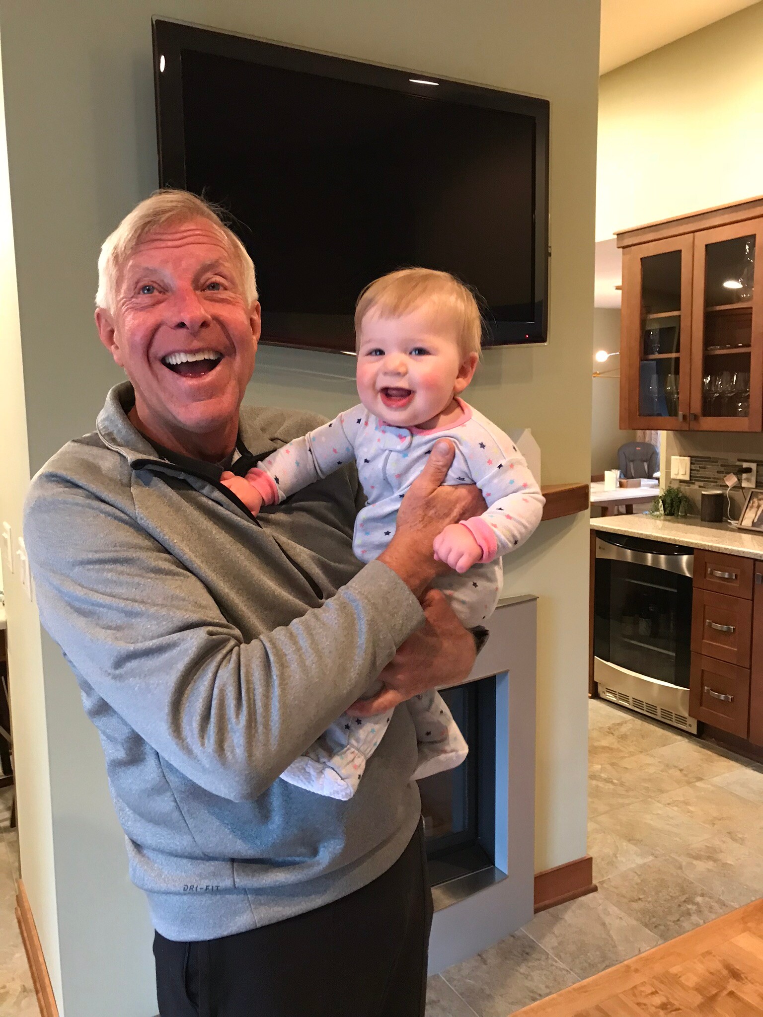 Older man in a gray hoodie laughing while cradling a smiling baby in a kitchen setting with a mounted TV behind them.