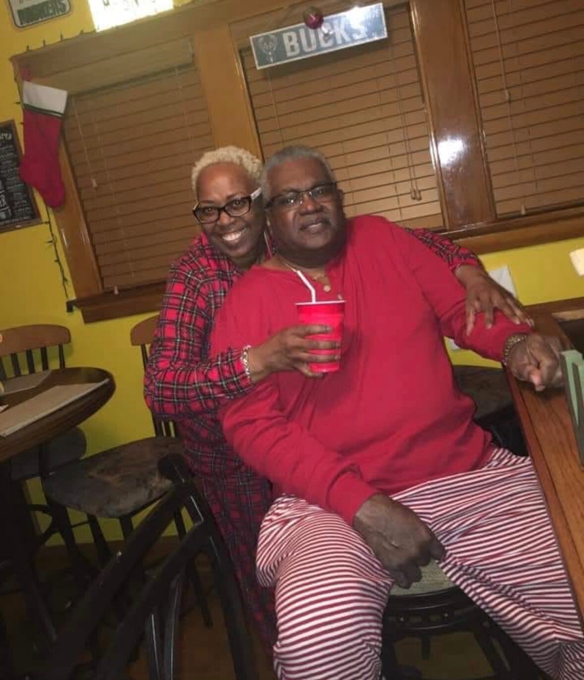 Two older adults pose together indoors; woman in plaid stands behind man in red, smiling and holding a red cup between them.