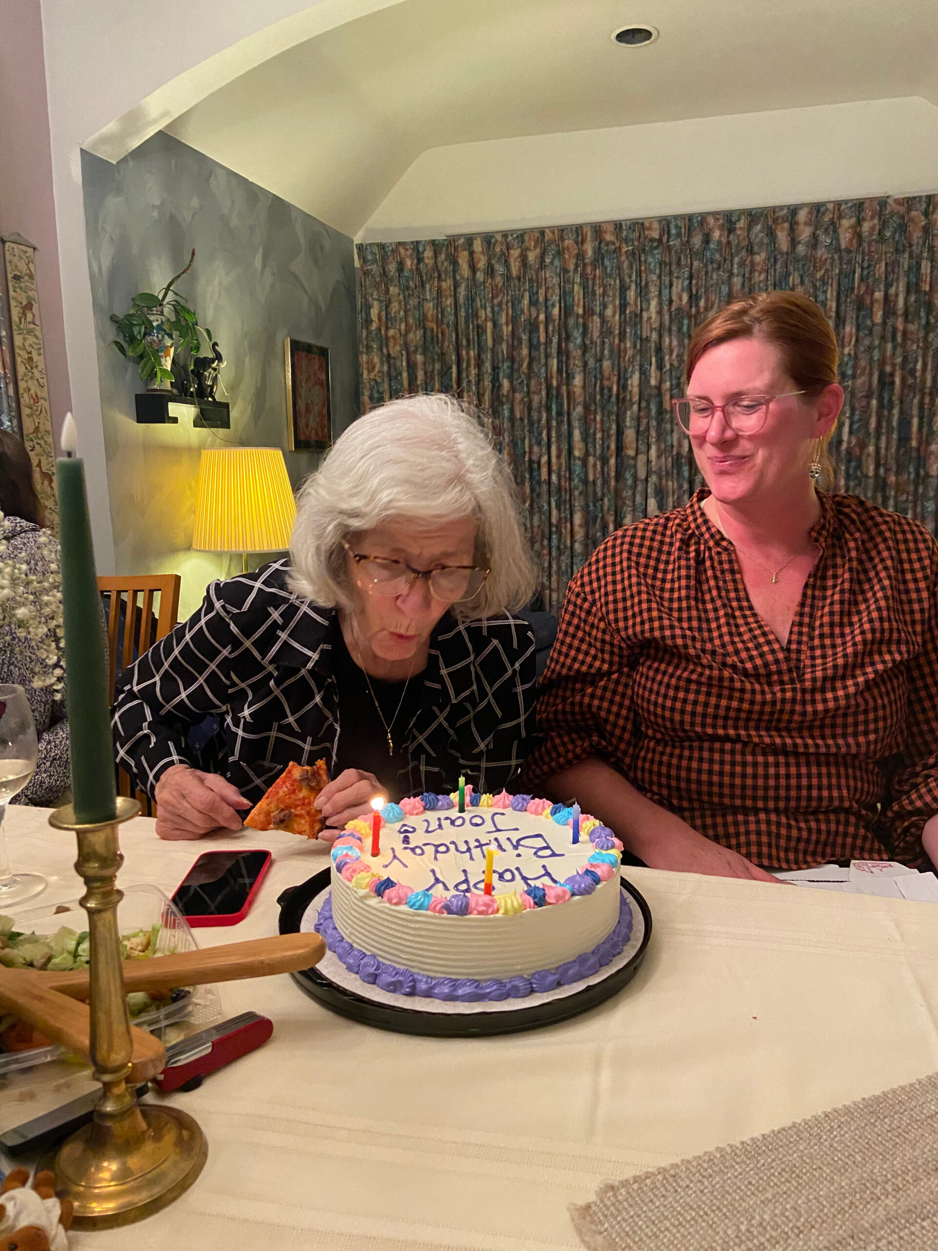 Two women at a birthday celebration; the older woman is cutting a slice of cake while the younger woman smiles nearby, with a frosted cake and candles on the table.