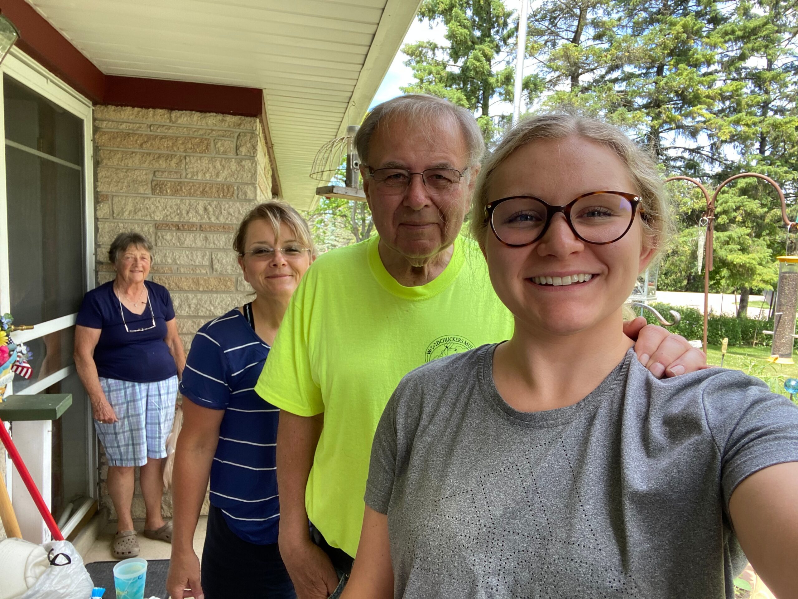 Young woman with glasses taking a selfie on a porch, smiling, with three older adults standing behind her.