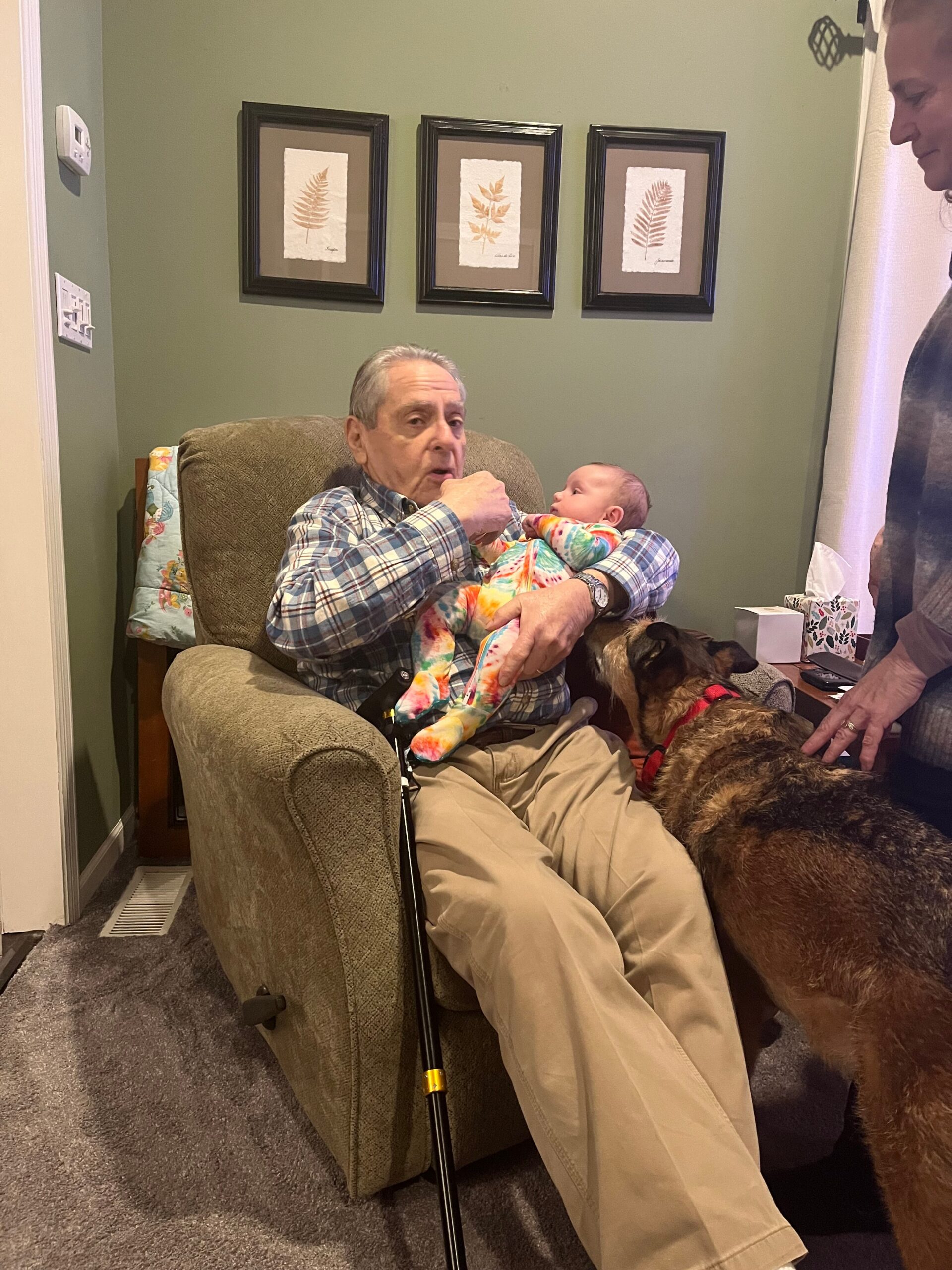 Older man sitting in an armchair cradles a newborn baby, with a dog at his feet in a cozy living room.