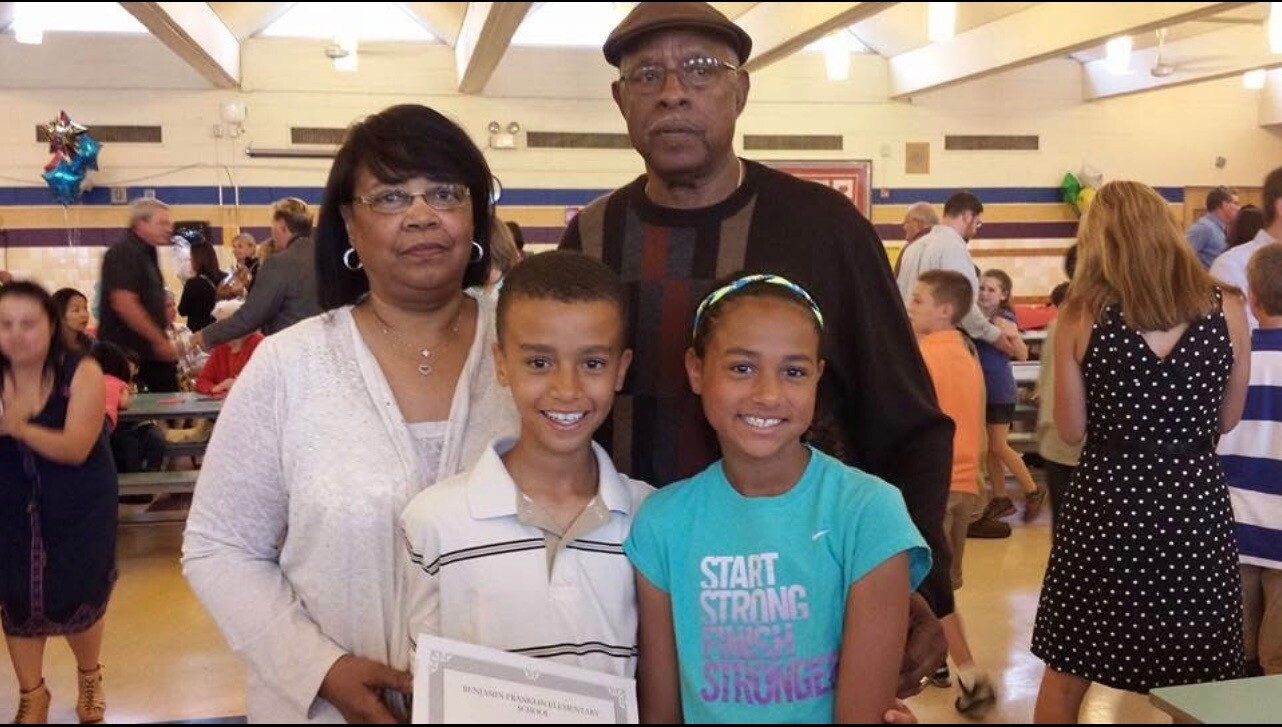 A family of four posing for a photo at a school event in a gymnasium, with two smiling children in front holding a certificate while their parents stand behind them.
