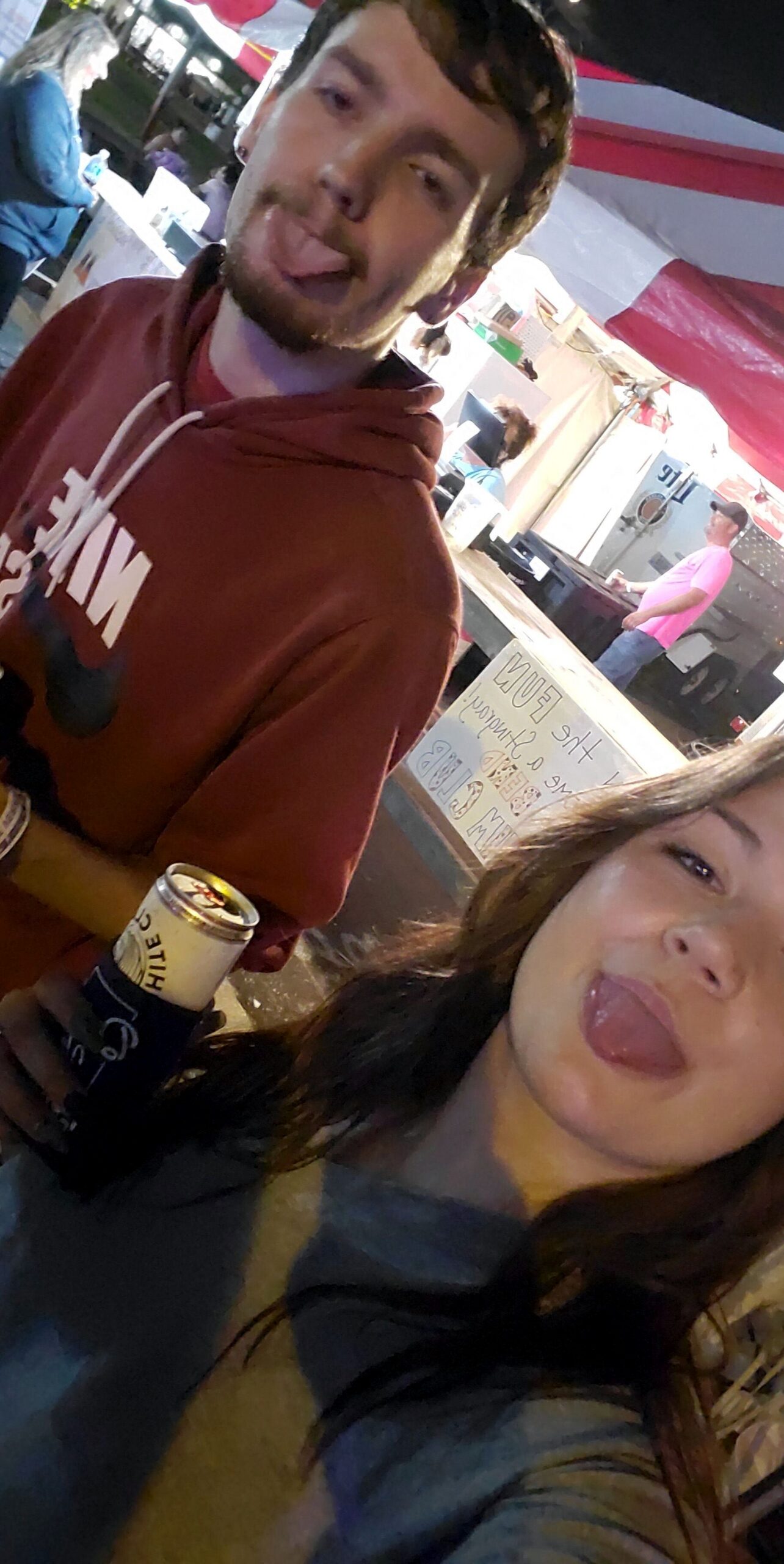 Two friends pose for a selfie at a night market or festival, both sticking out their tongues; man in a maroon hoodie holds a can.