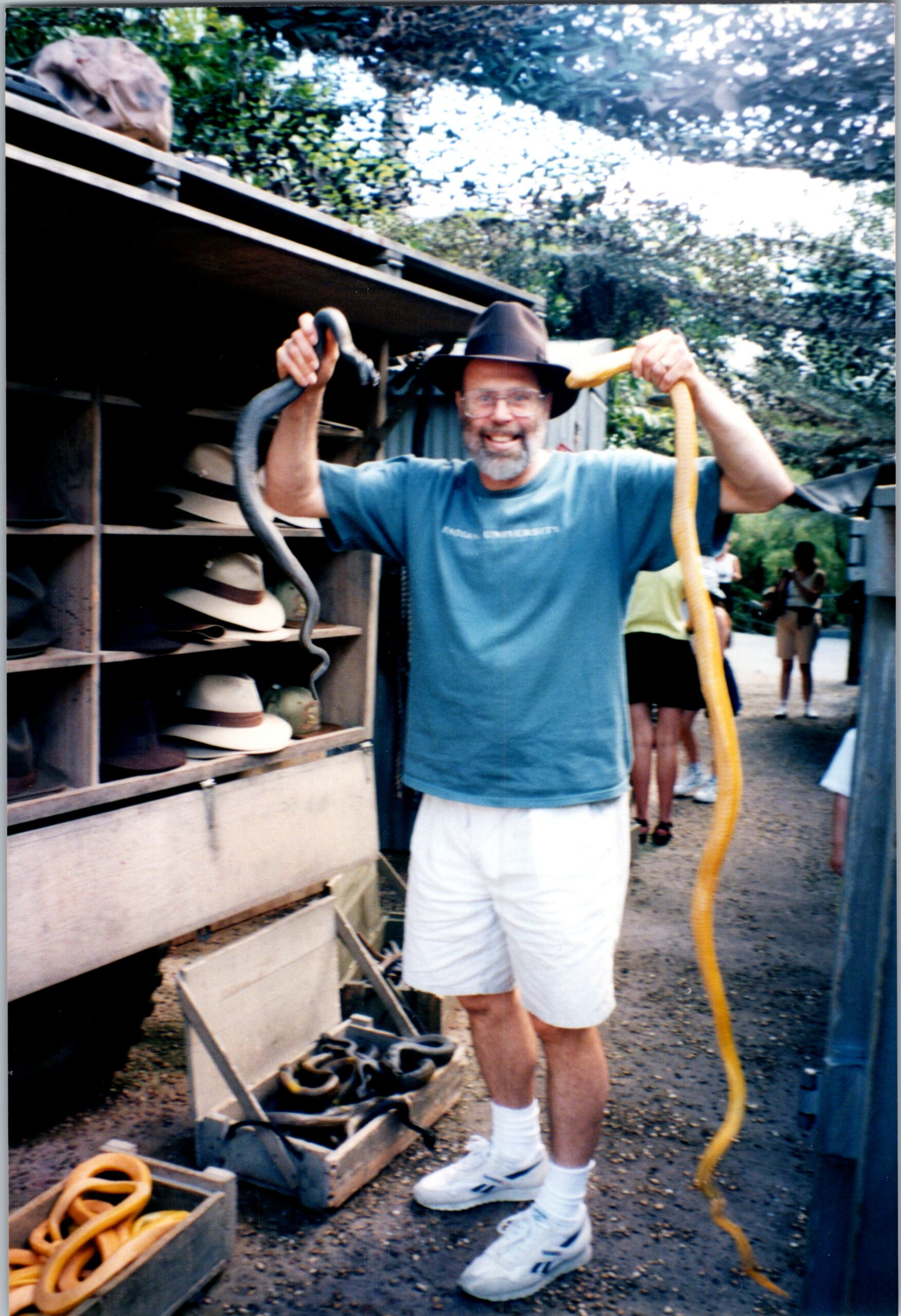 Man in a blue shirt and hat proudly holds two snakes, a black one and a yellow one, outdoors beside a hat display.