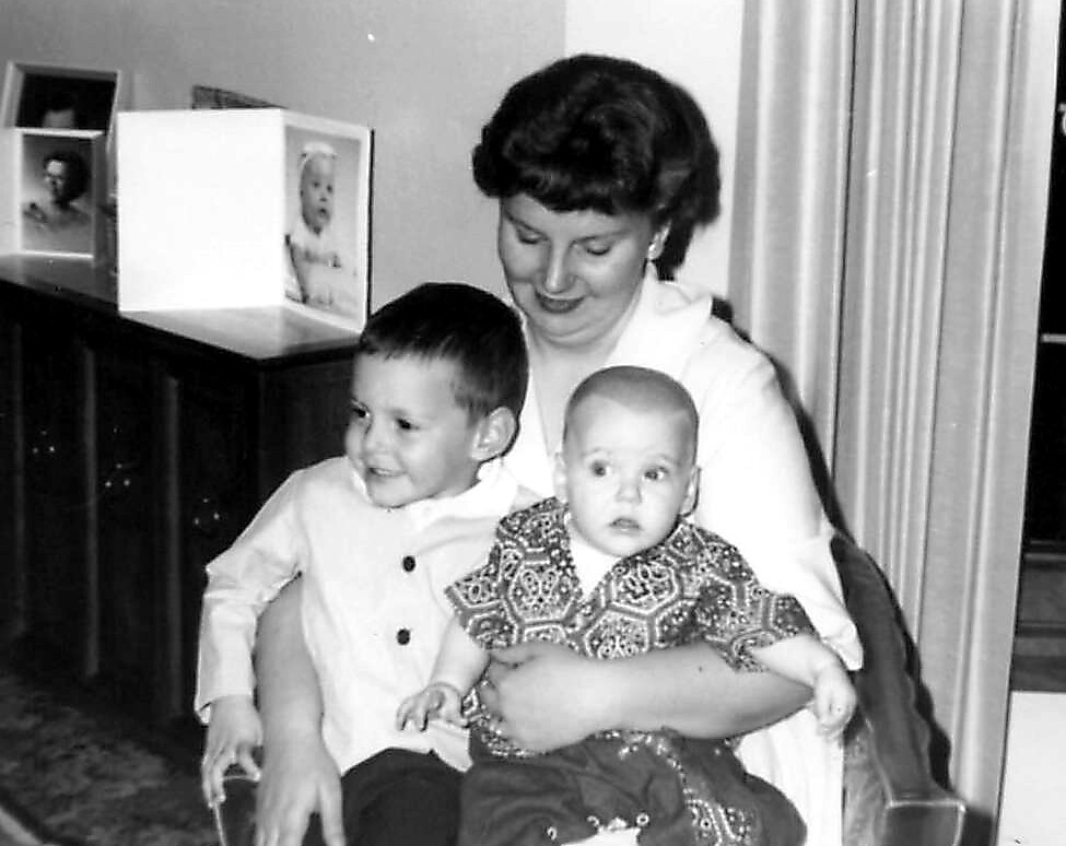 Mother seated with two young children: a toddler on her left and a baby on her lap, in a home setting and smiling down at them