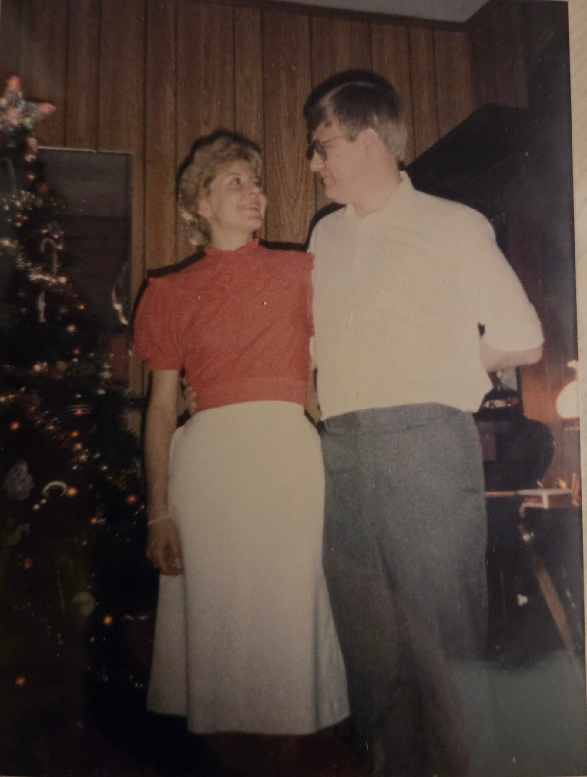 Smiling couple posing indoors near a Christmas tree, looking at each other.