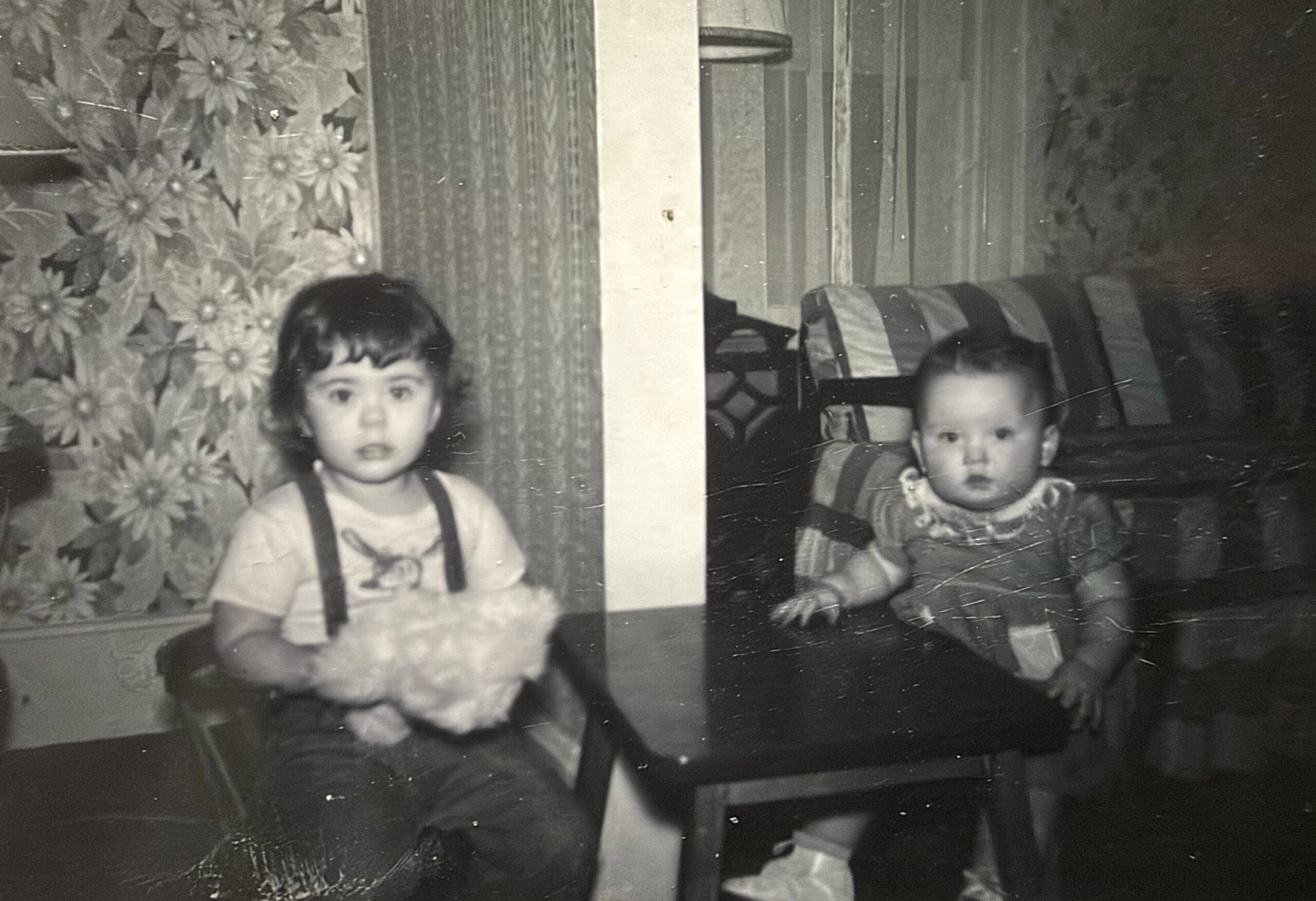 Two young children sit at a table indoors; left child wears suspenders and holds a bouquet, right child sits in a high chair, vintage room with floral wallpaper in background.