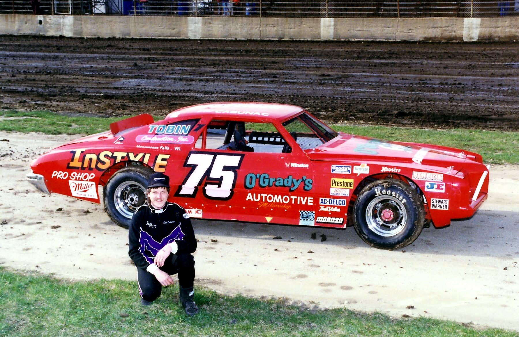 Red race car with the number 75 and sponsor decals on a dirt track; a racer kneels beside it in racing gear.