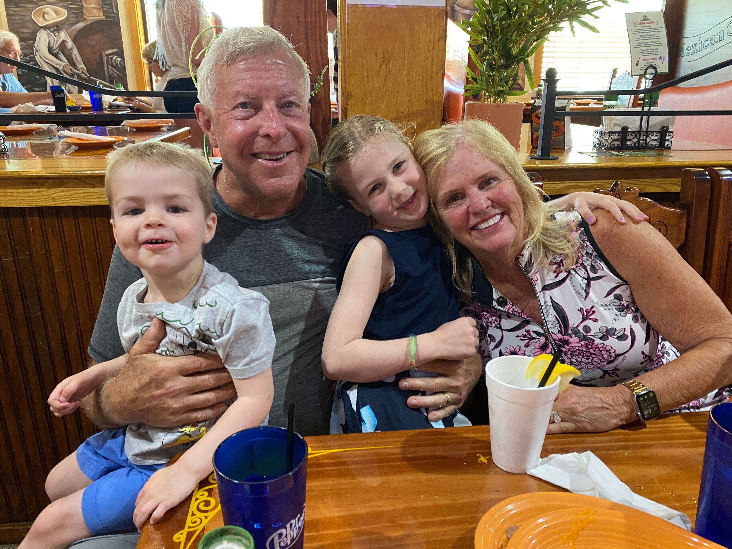 A smiling family posing together at a restaurant: grandfather with toddler, girl and woman leaning in, drinks on the wooden table in front of them.