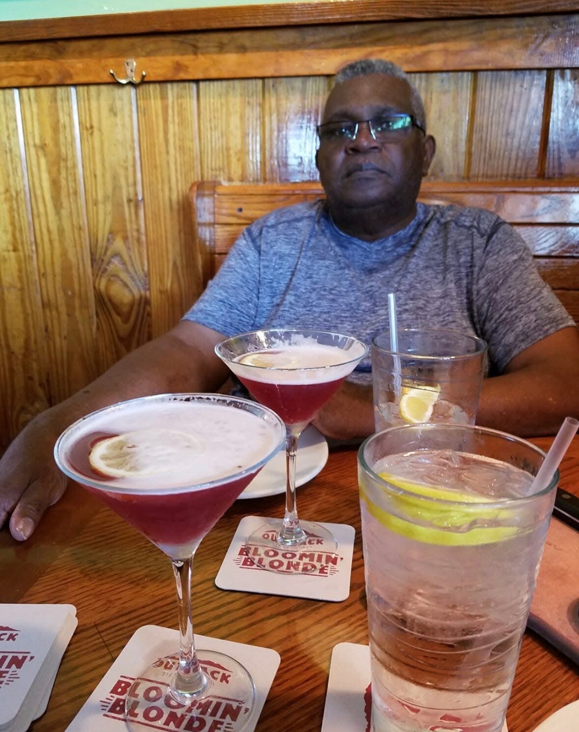 Man wearing glasses sits at a wooden booth with three drinks on the table: two pink cocktails in martini glasses and a lemon-water tall glass in front of him.