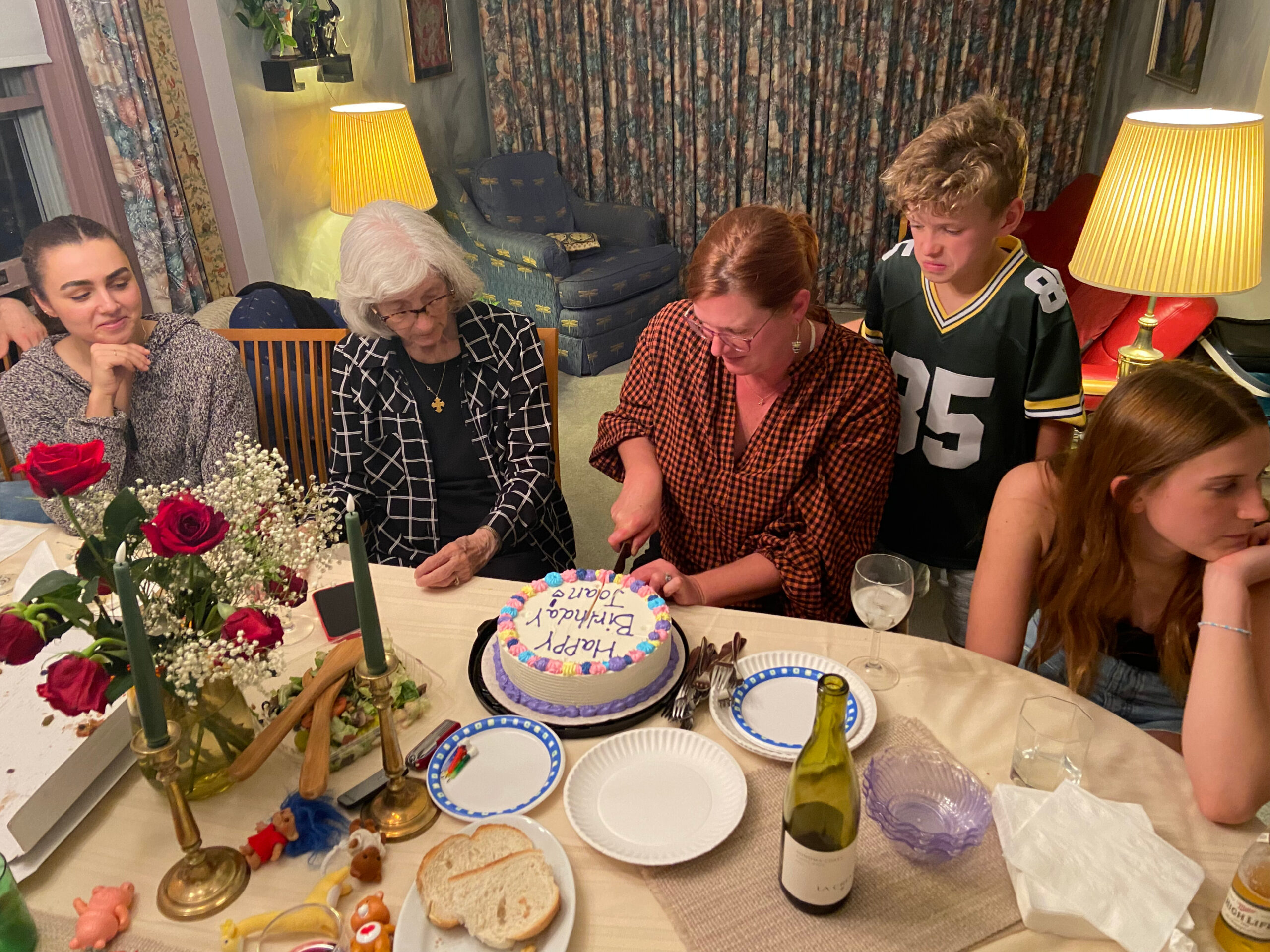 Group of five people around a table cutting a birthday cake, with candles, flowers, and wine in a cozy living room setting.