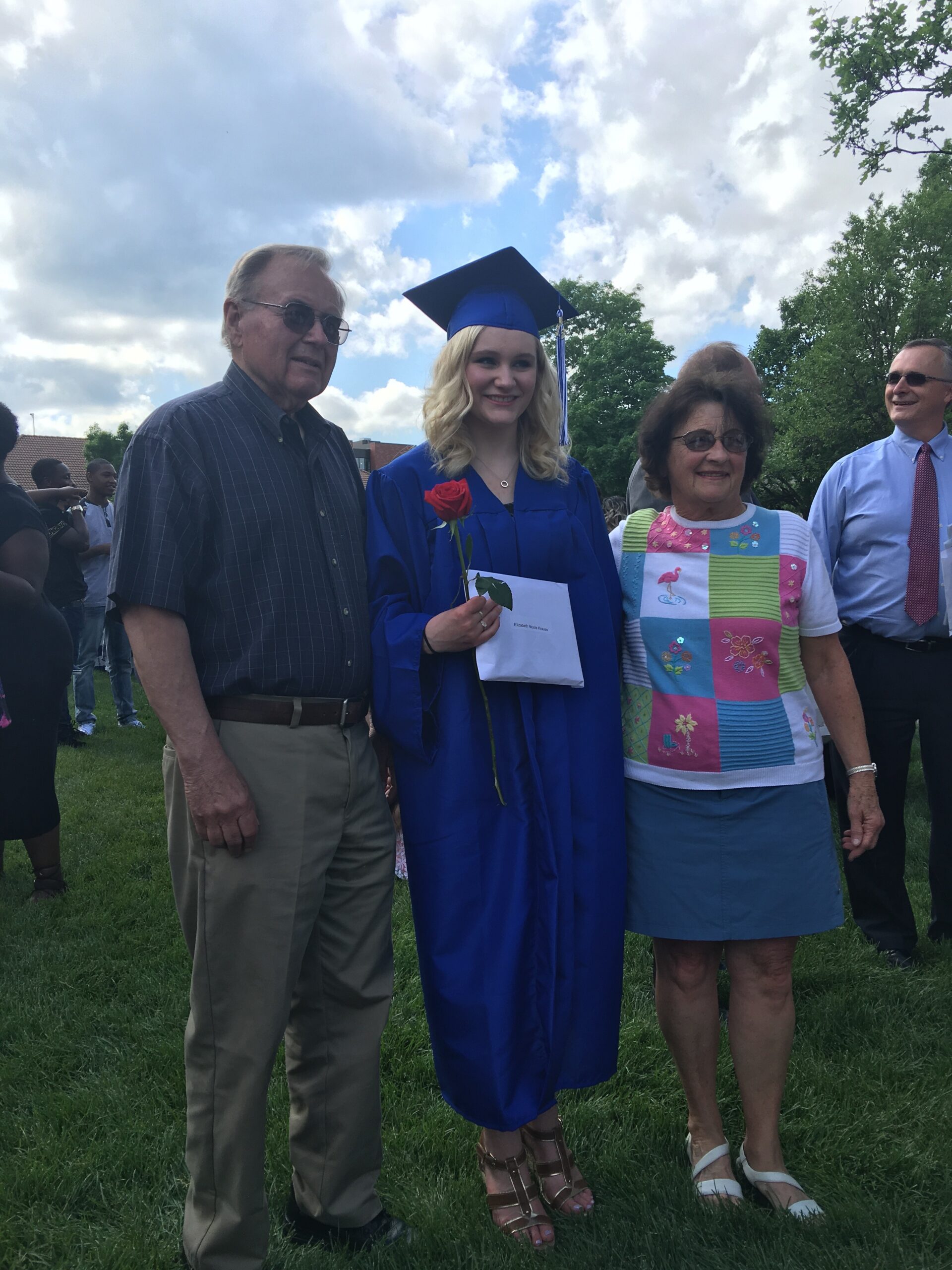 Graduate in blue cap and gown holding a red rose and diploma envelope, flanked by smiling adults outdoors on grass.