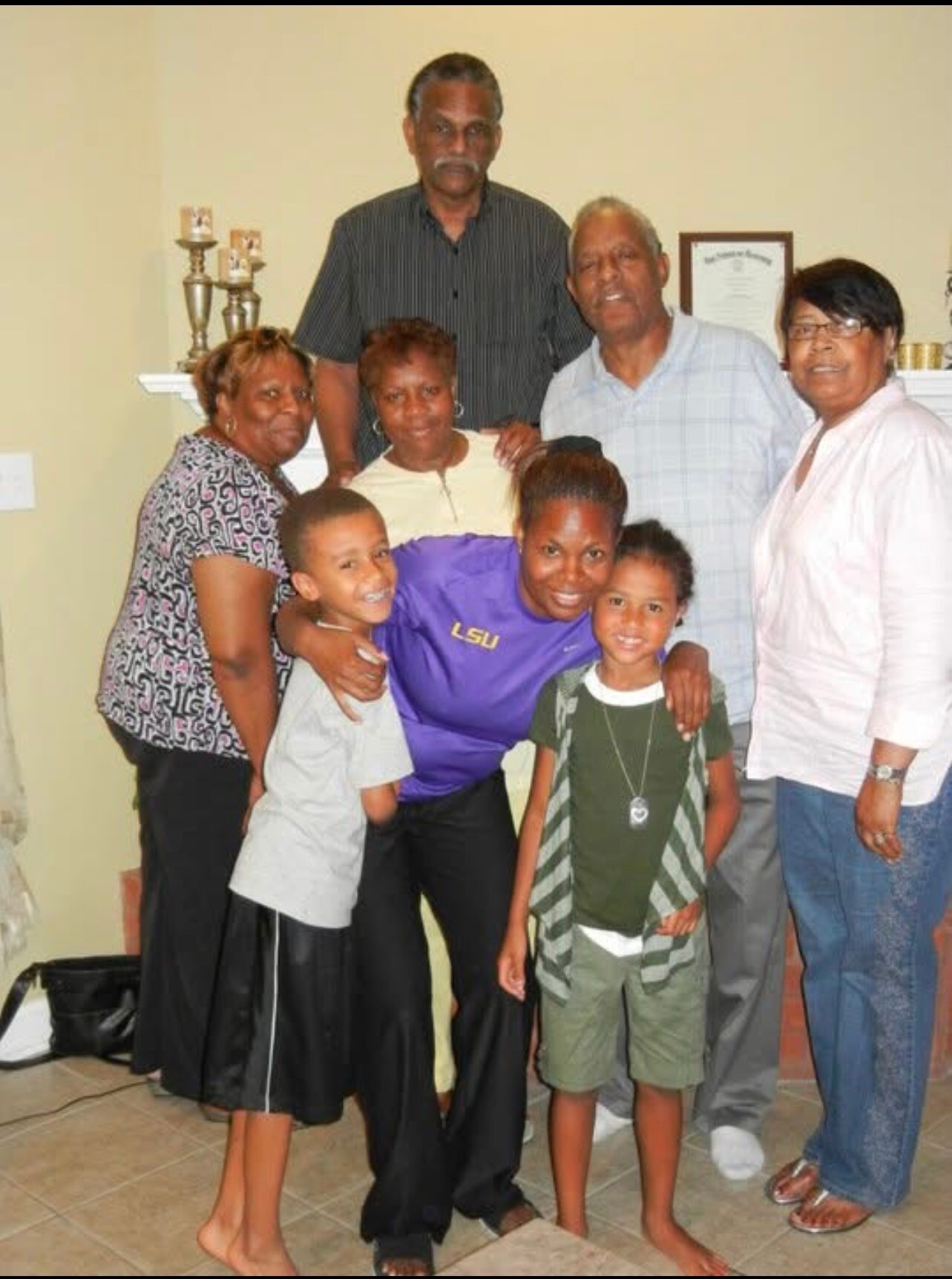 A multi-generation family group stands together in a living room, smiling for the photo, with a shelf and trophies in the background.