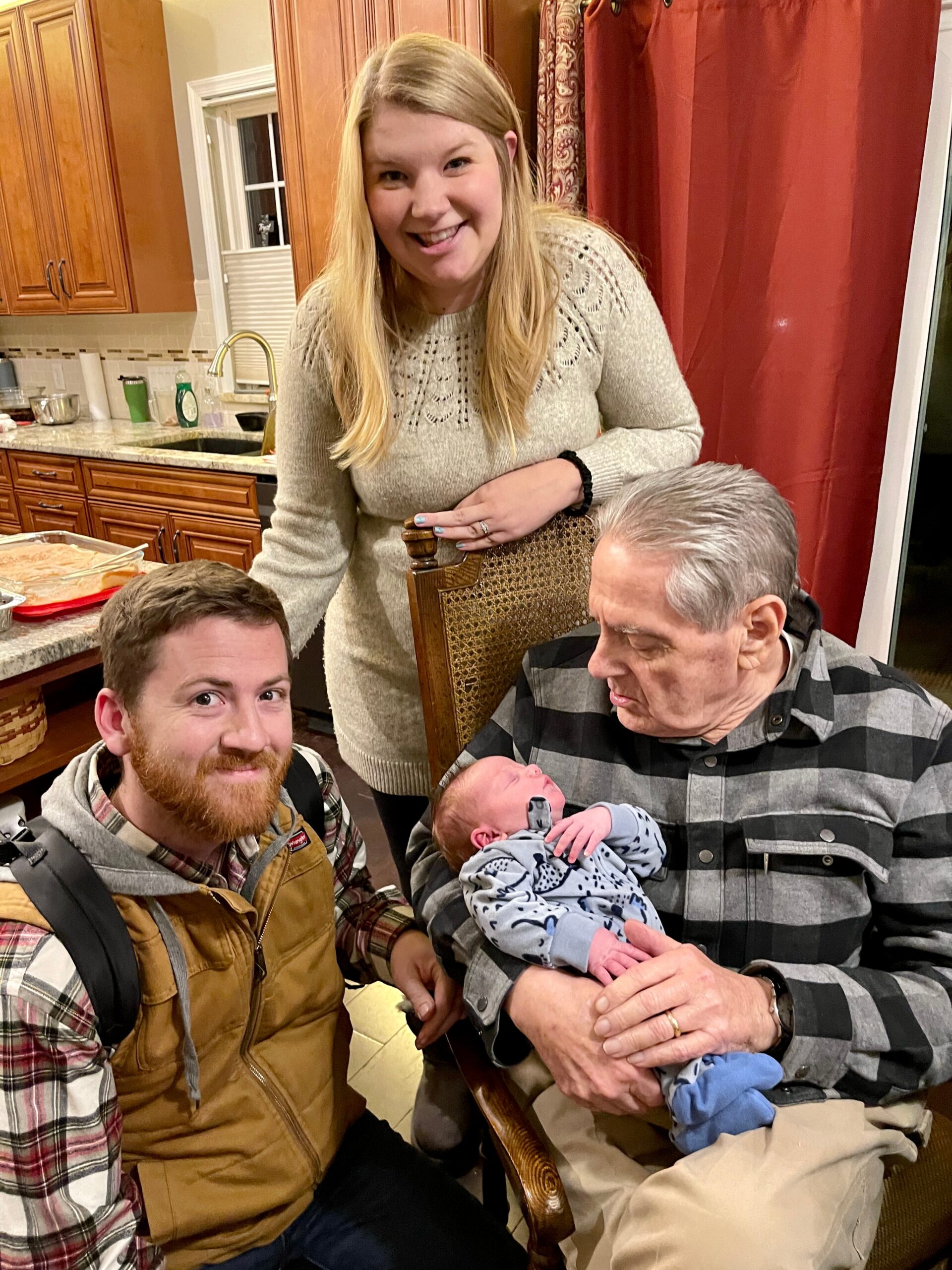 Smiling woman stands behind two men at a kitchen table, one holding a newborn baby wrapped in a striped blanket.
