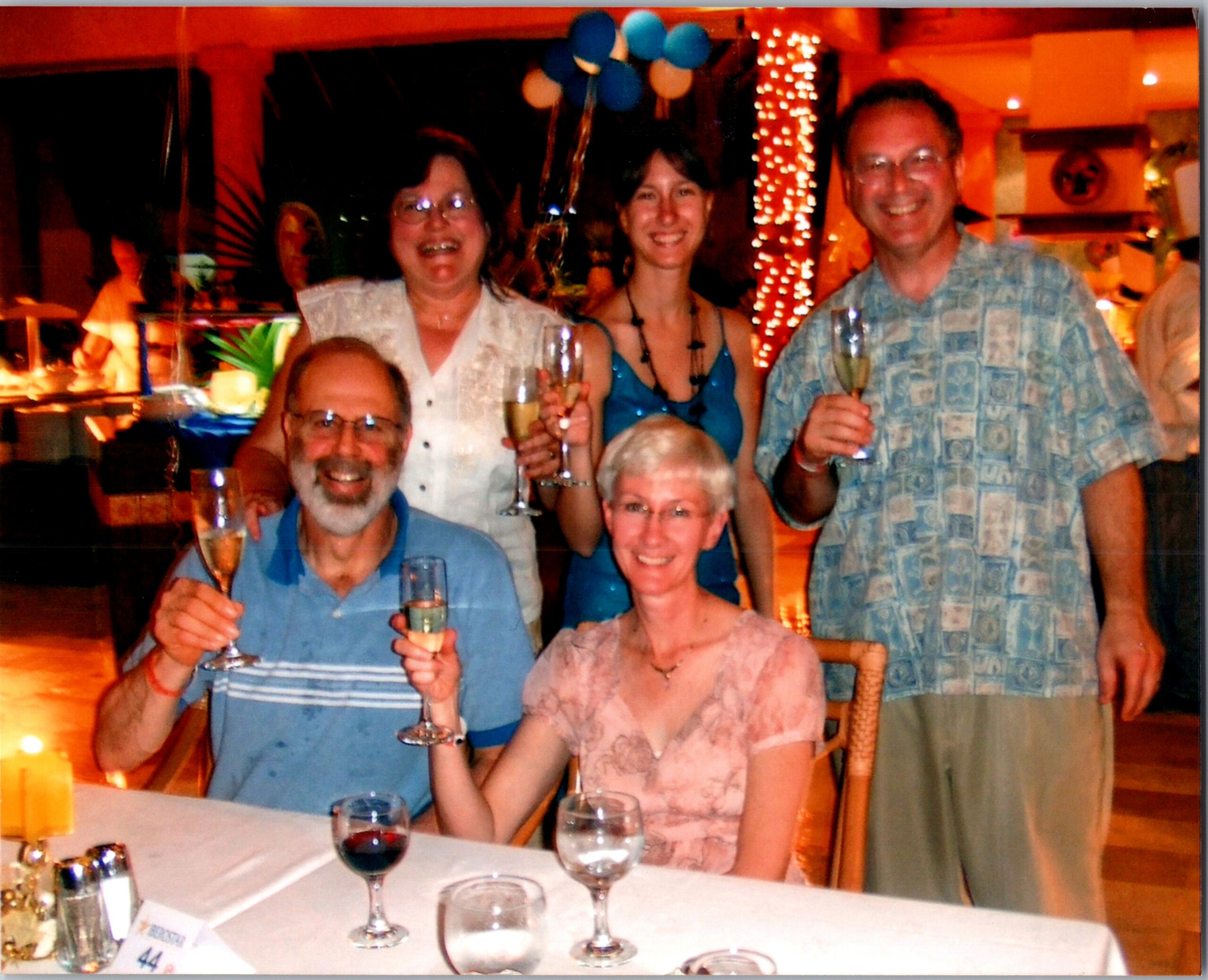 Group of smiling adults at a celebration, raising champagne glasses around a table with candles and drinks in a warm, festive setting.