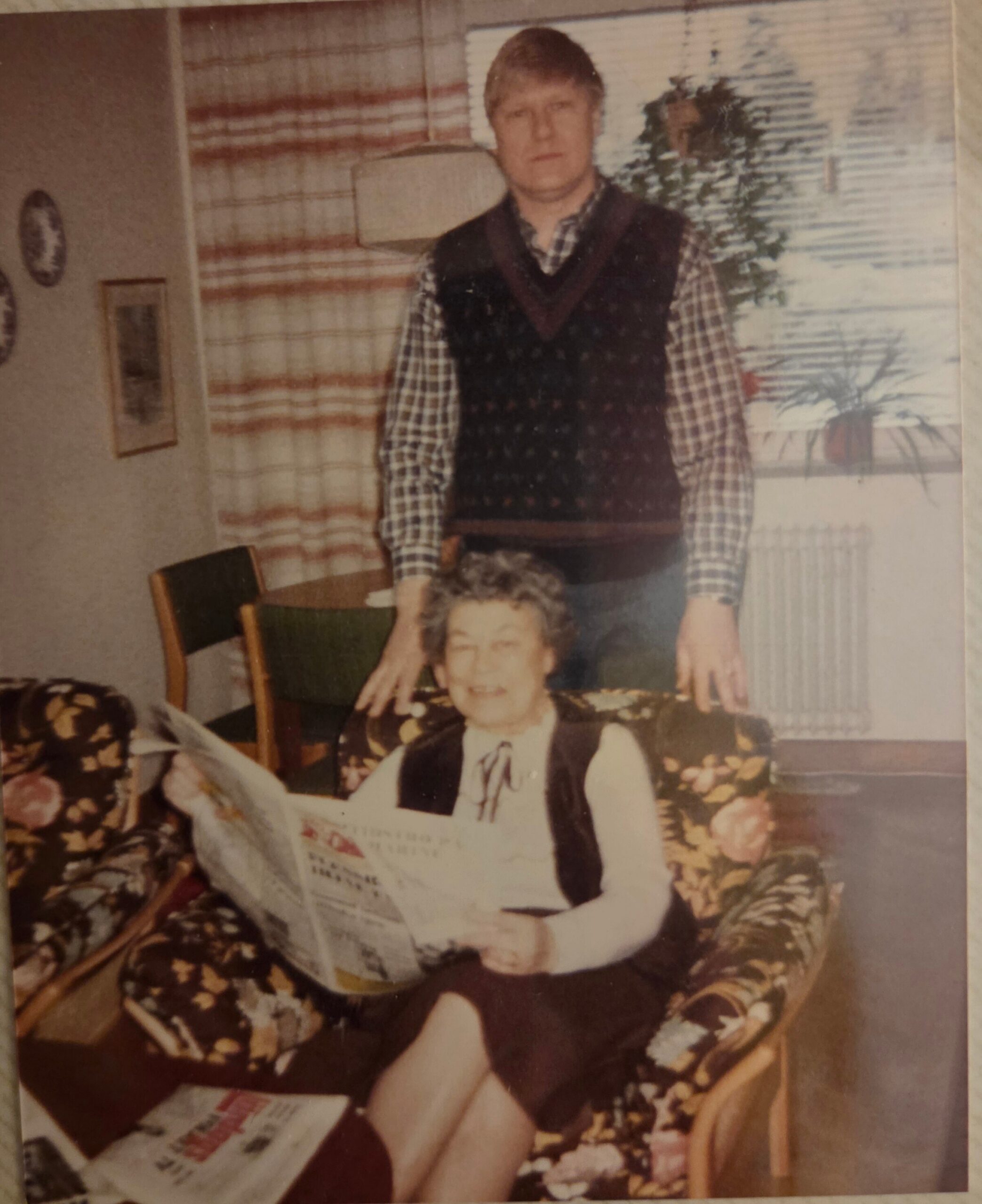 A man in a plaid shirt and dark sweater vest stands behind an elderly woman who is seated and reading a newspaper in a floral living room.
