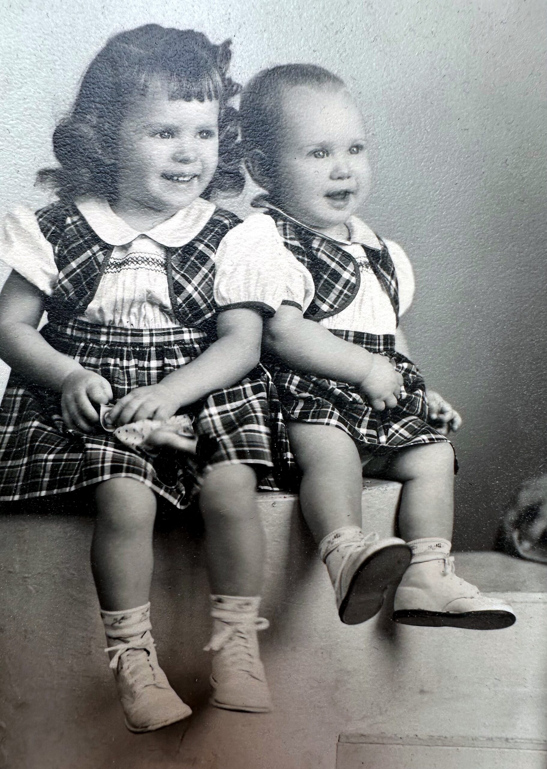 Two young children in matching plaid dresses sit side by side, smiling for a portrait.