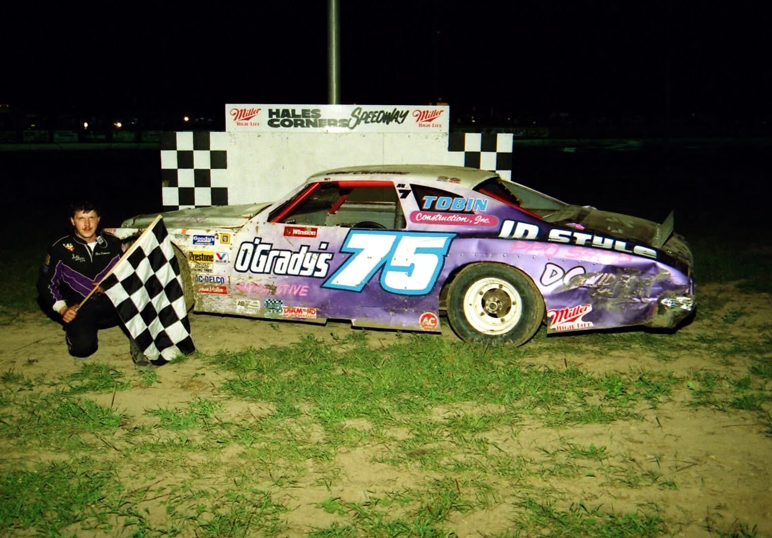 Race car number 75 with purple/white livery and sponsor logos, driver kneeling with a checkered flag at night on a dirt track finish line.