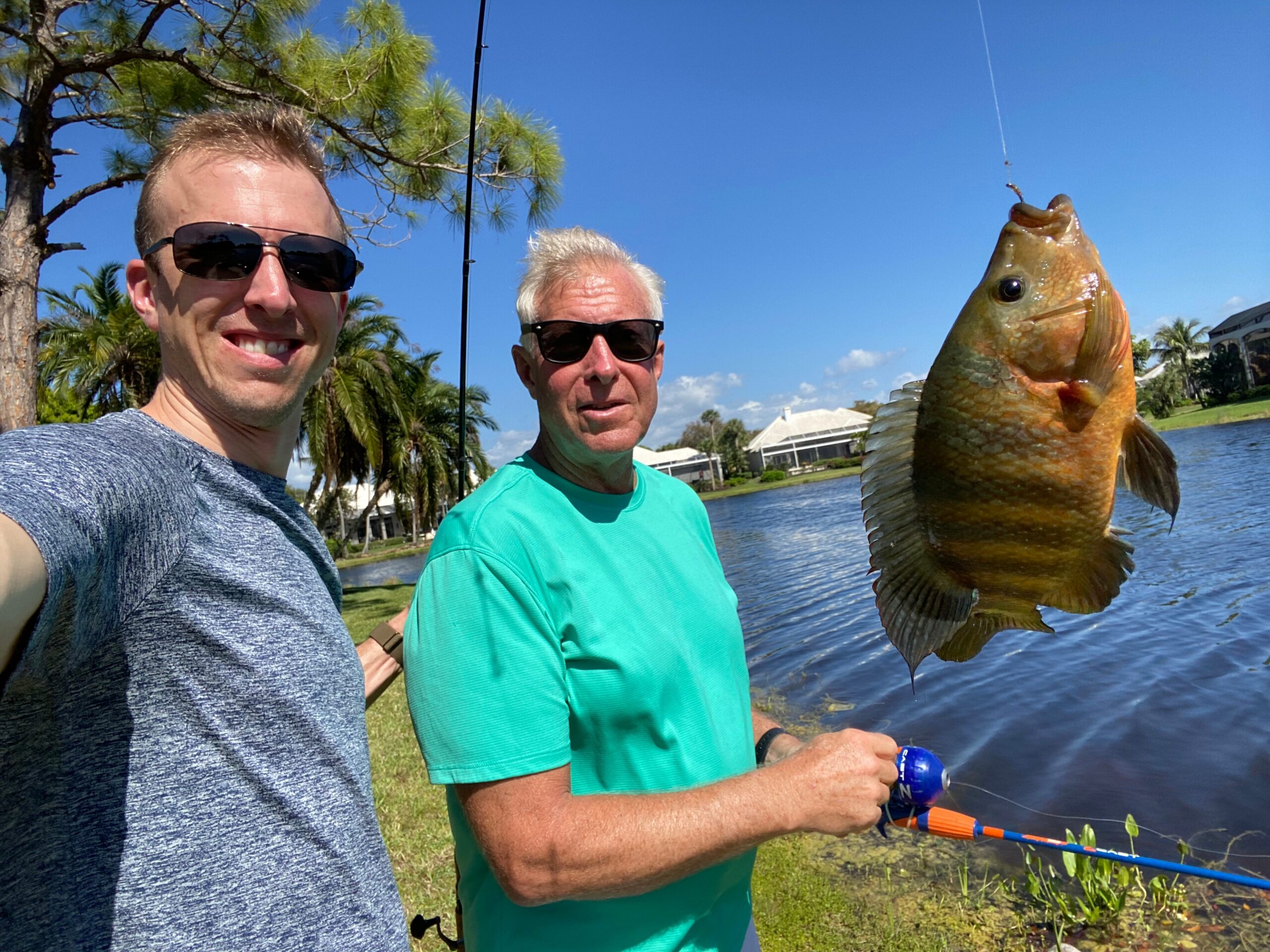 Two men stand by a lake on a sunny day, posing with a large fish they caught.