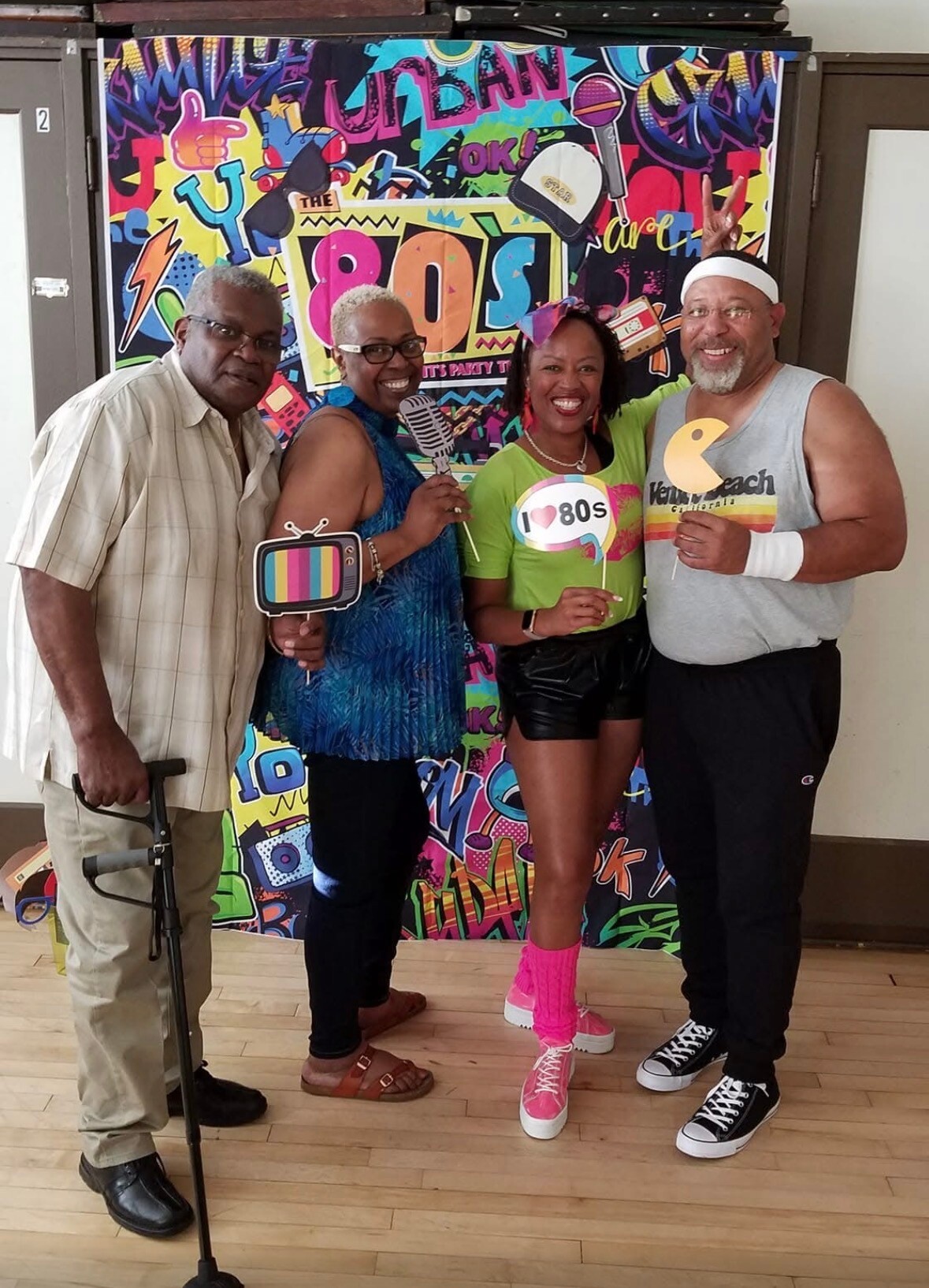 Four adults posing with retro props in front of a colorful 1980s-themed backdrop; they hold a TV prop, a microphone, a heart/80s sign, and peace sign gesture.