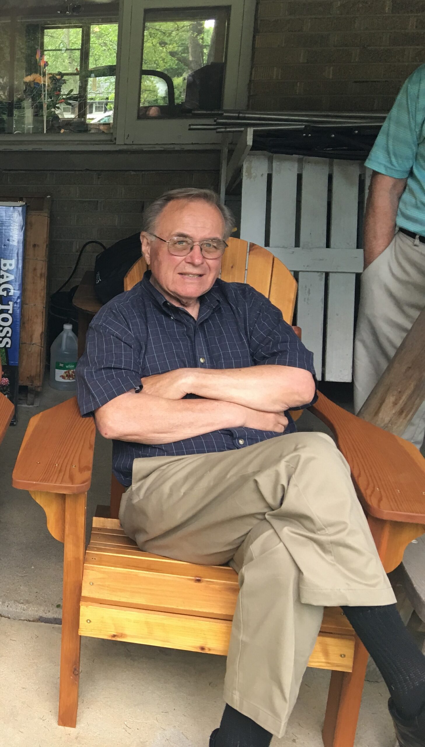 Older man with glasses sits in a wooden chair on a porch, arms folded, wearing a dark blue plaid shirt and khaki pants; another person stands nearby.