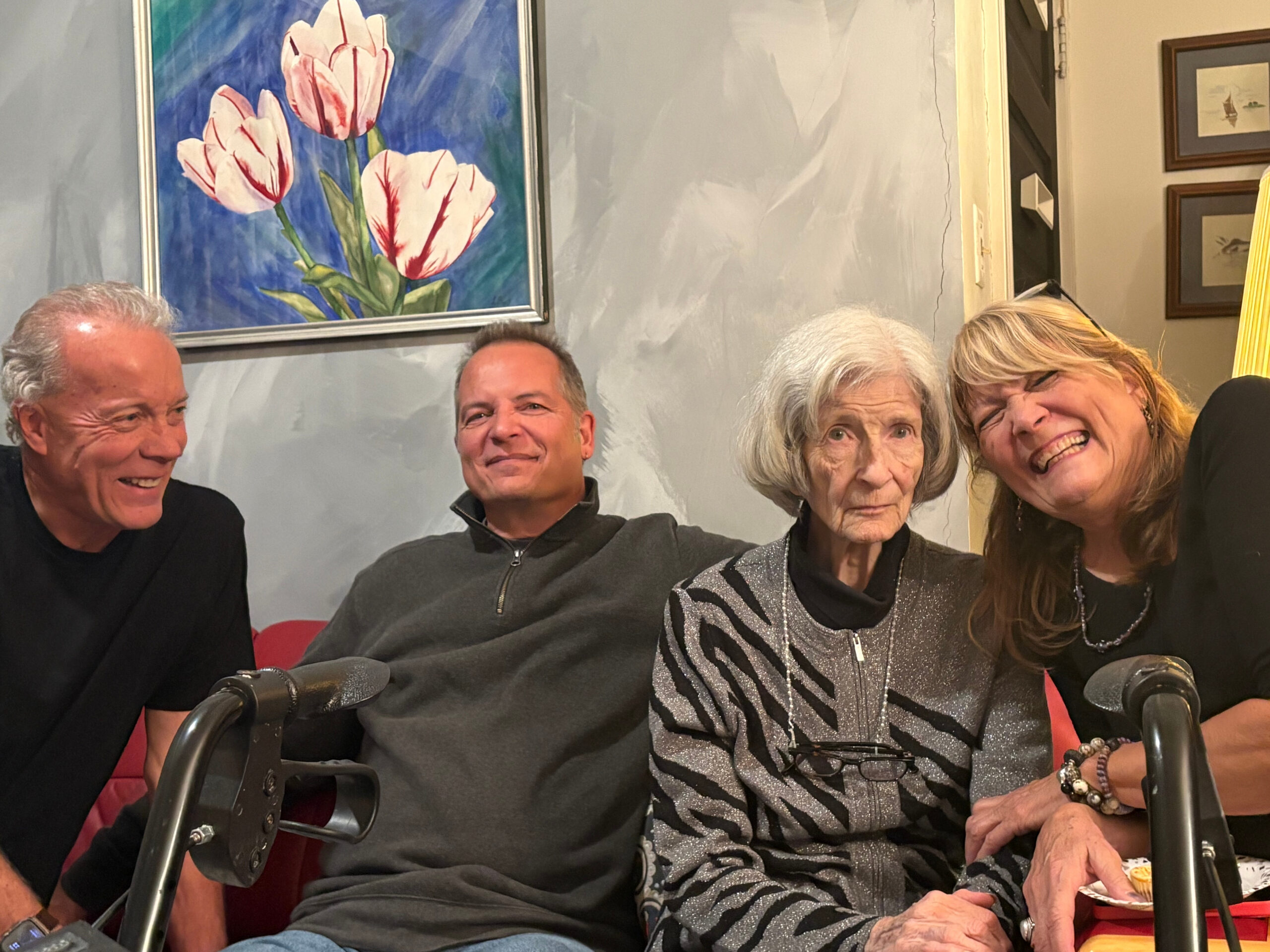 Four adults sitting on a couch indoors, smiling for a group photo; a painting of pink tulips hangs on the wall behind them.