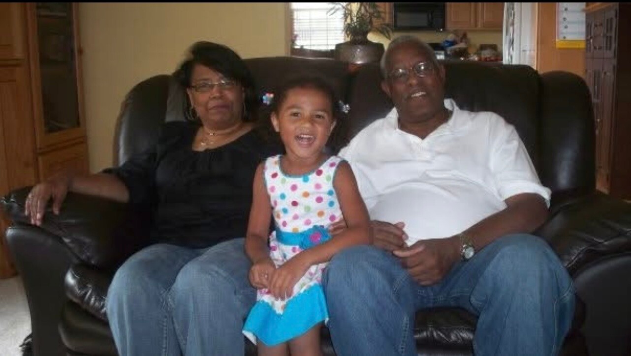 Young girl in a colorful polka‑dot dress sits between smiling grandparents on a brown leather sofa in a living room.