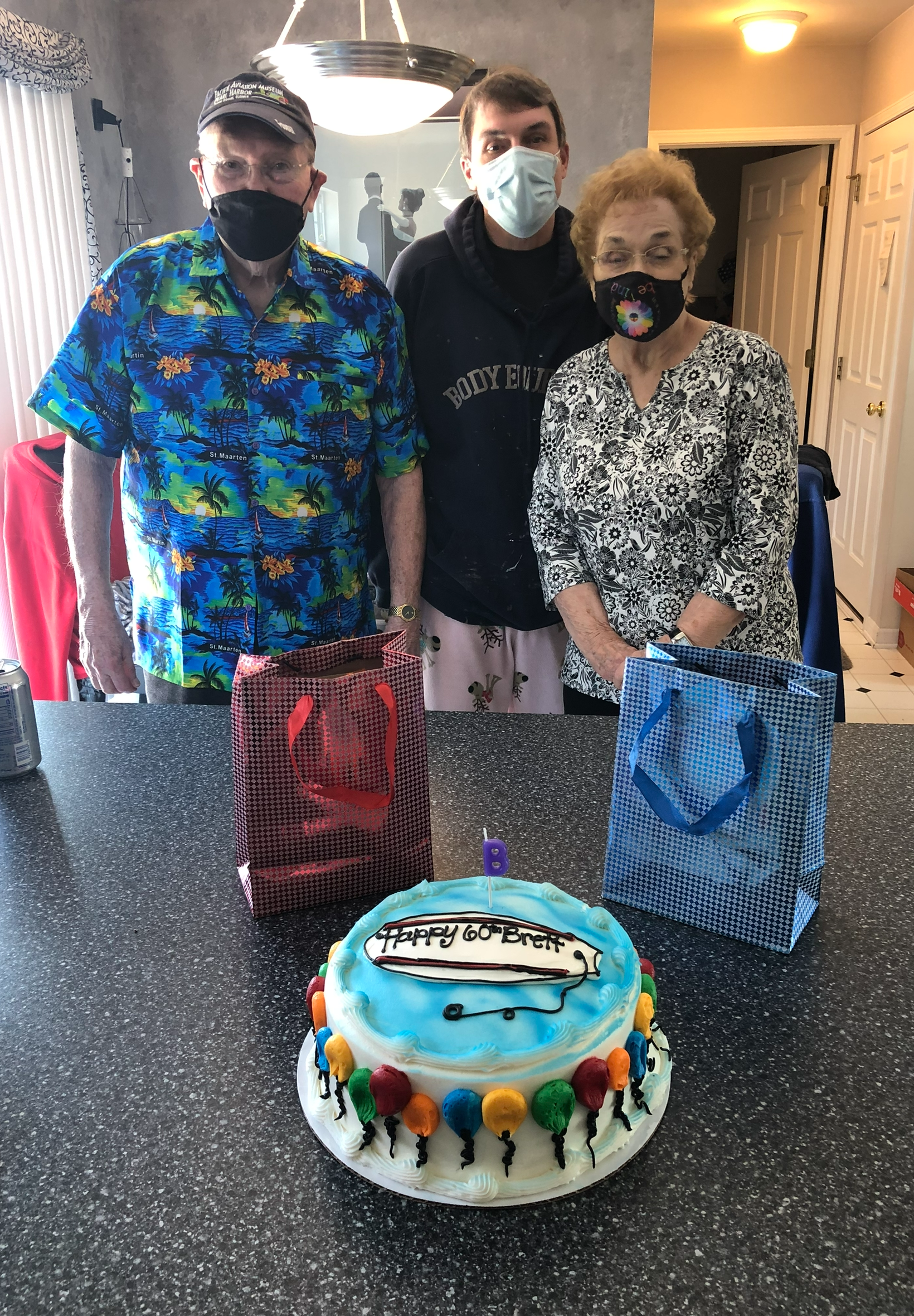 Three people wearing masks pose behind a decorated birthday cake and gift bags on a kitchen counter.