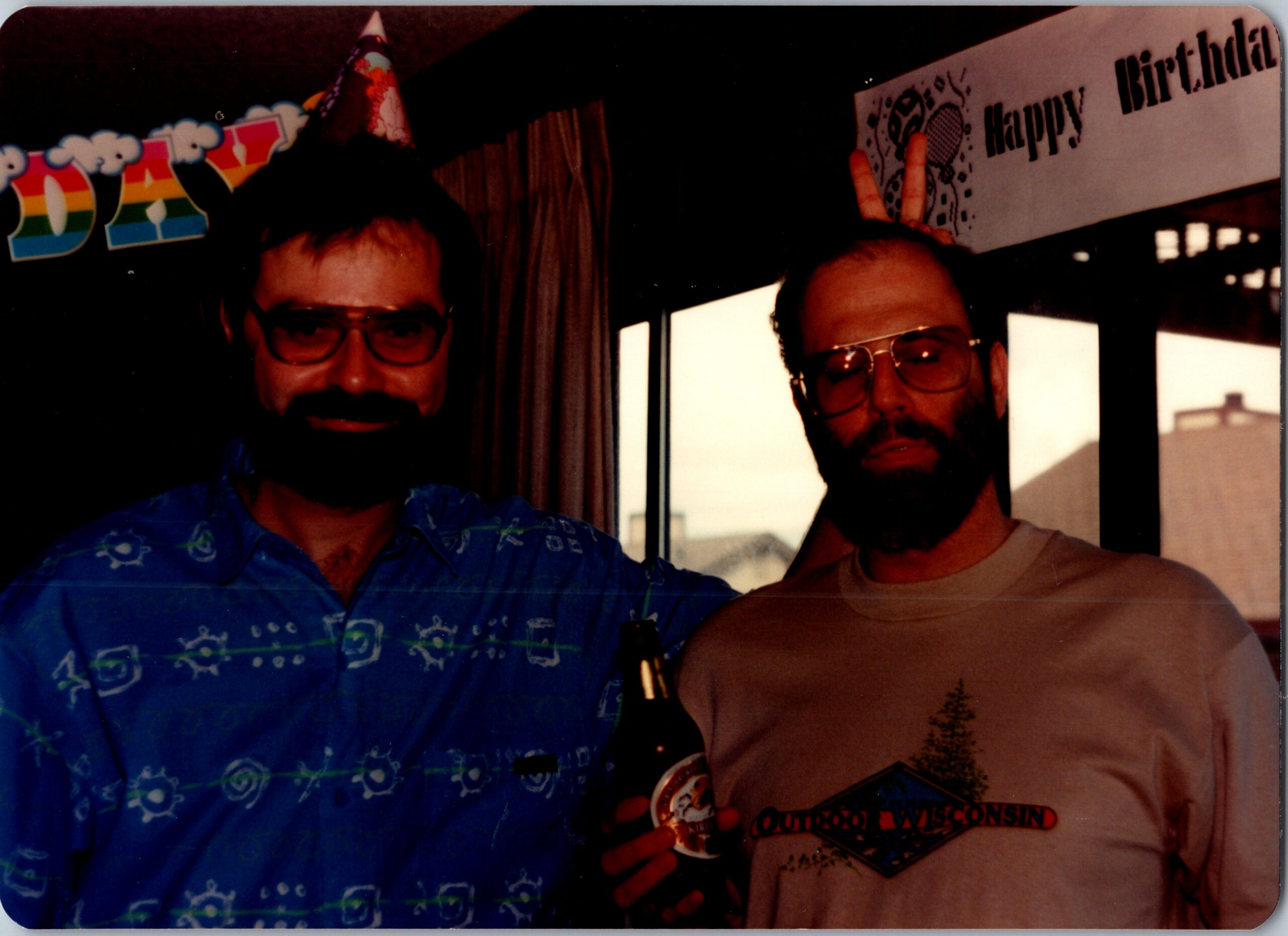 Two men at a birthday party wearing party hats; one holds a beer bottle, with a 'Happy Birthday' banner in the background between them.