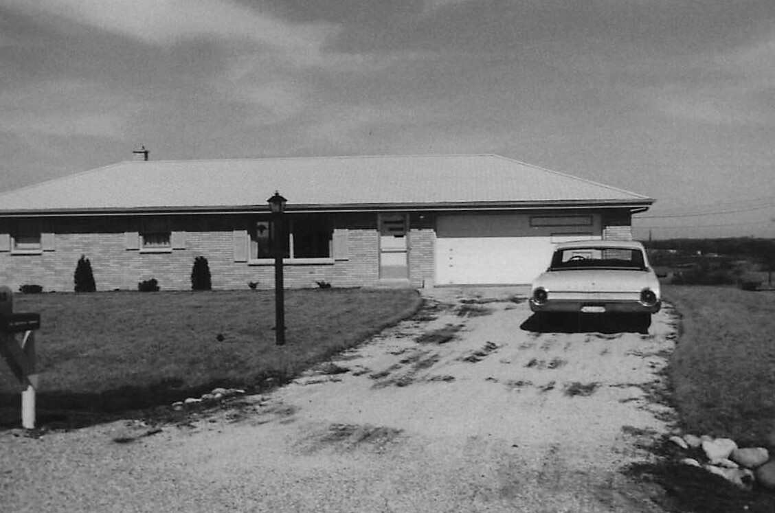 Front view of a single-story brick house with a gravel driveway and a vintage car parked in front.