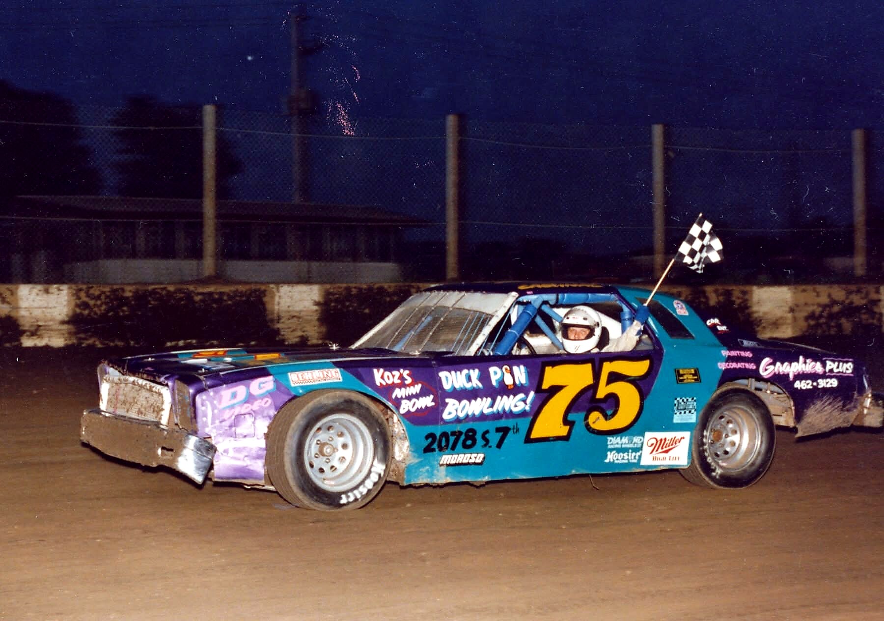 Nighttime dirt-track race car #75 with a driver in a white helmet pilot­ing along the track, a black-and-white checkered flag mounted on the car's rear.