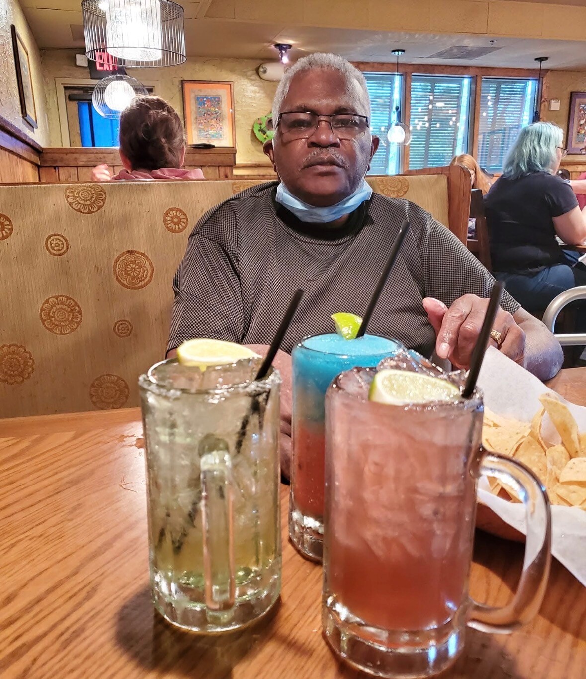 A smiling older man with glasses sits at a booth in a restaurant, wearing a gray shirt and a blue face mask pulled under his chin, with three colorful cocktails in front of him on the table.