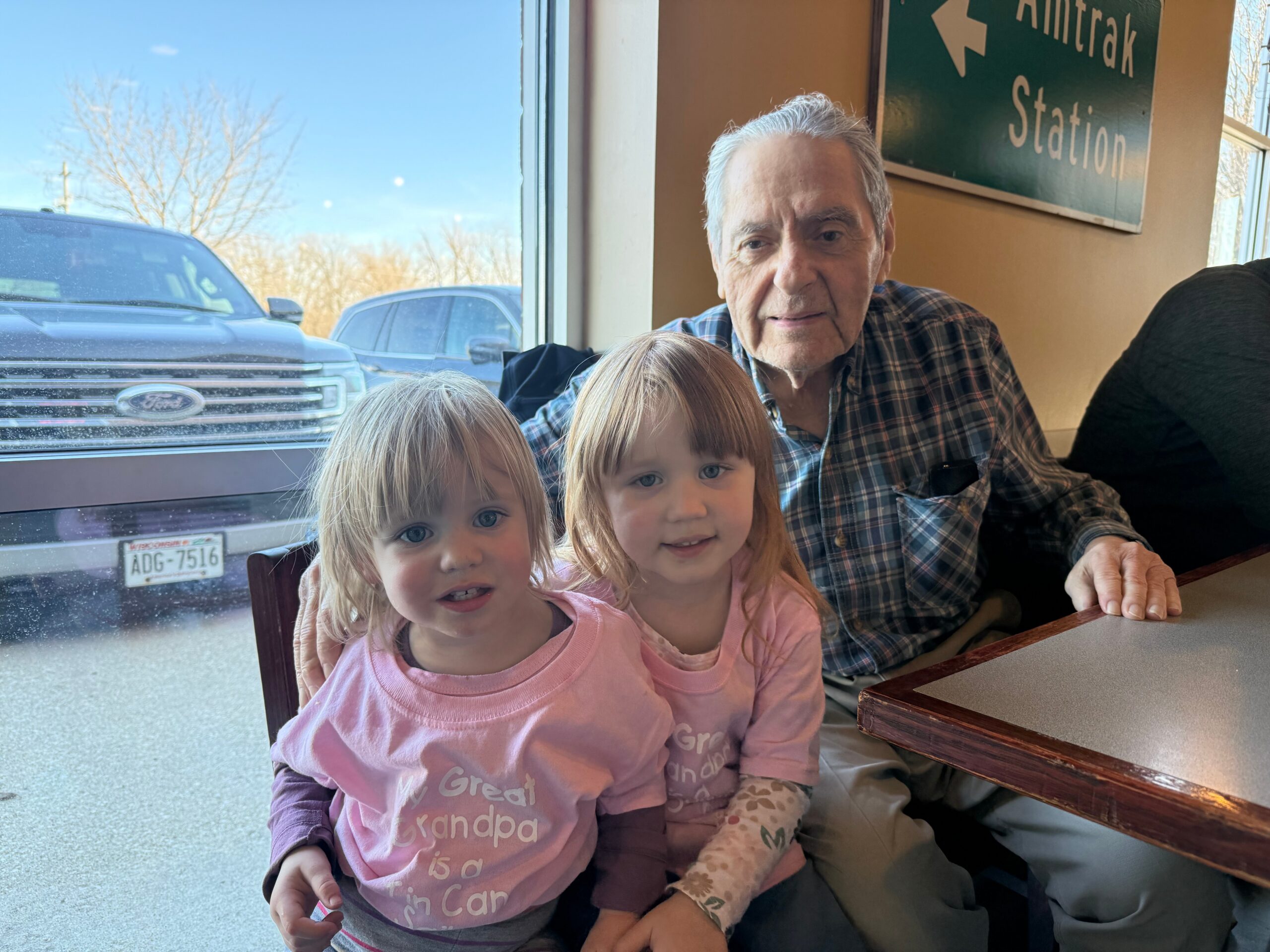 Two young girls sit on laps with an older man at a booth by a window in a diner; a green Amtrak Station sign is on the wall behind them.