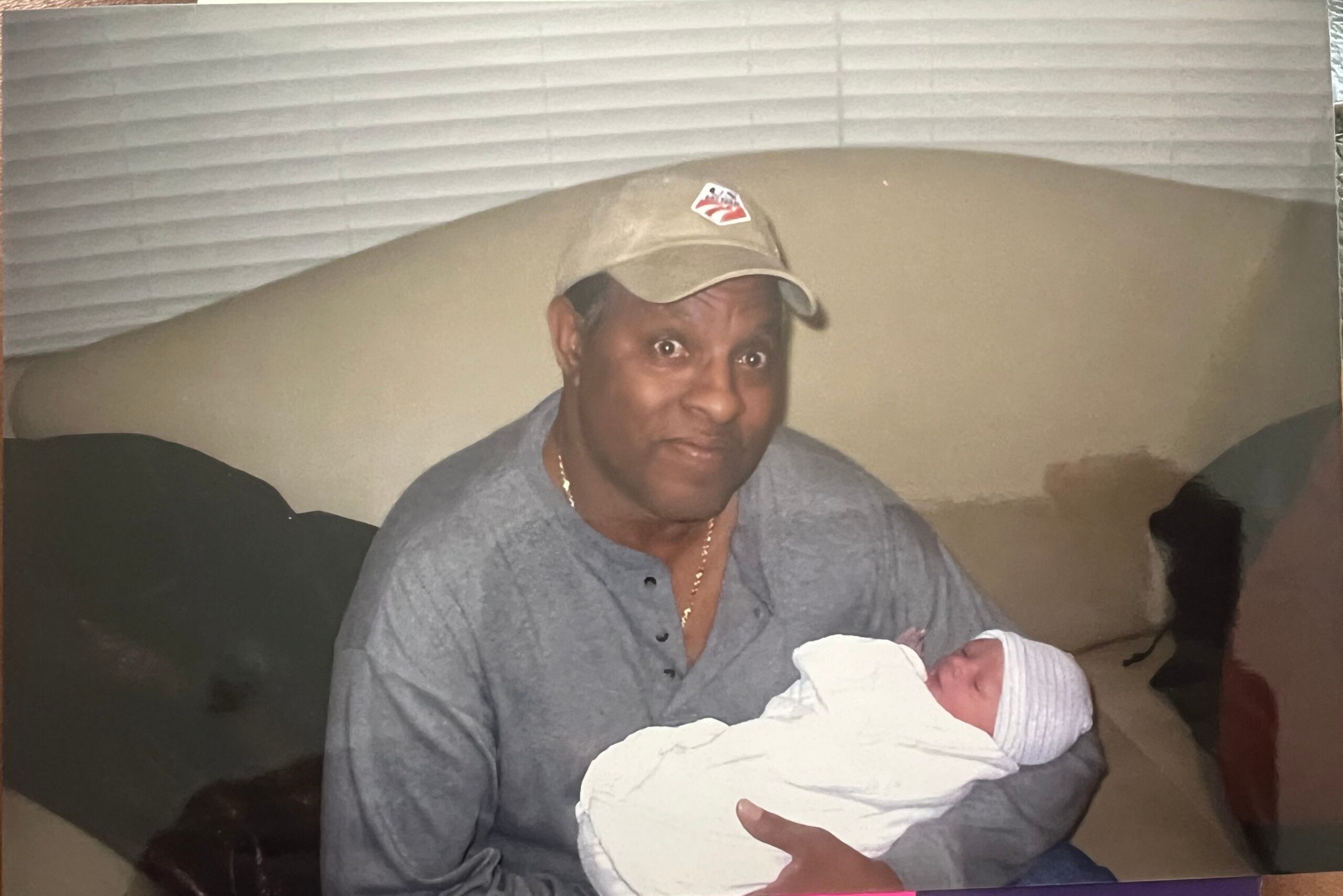 Man in a gray shirt and cap cradling a newborn wrapped in a white blanket, smiling at the baby in a warm indoor setting.