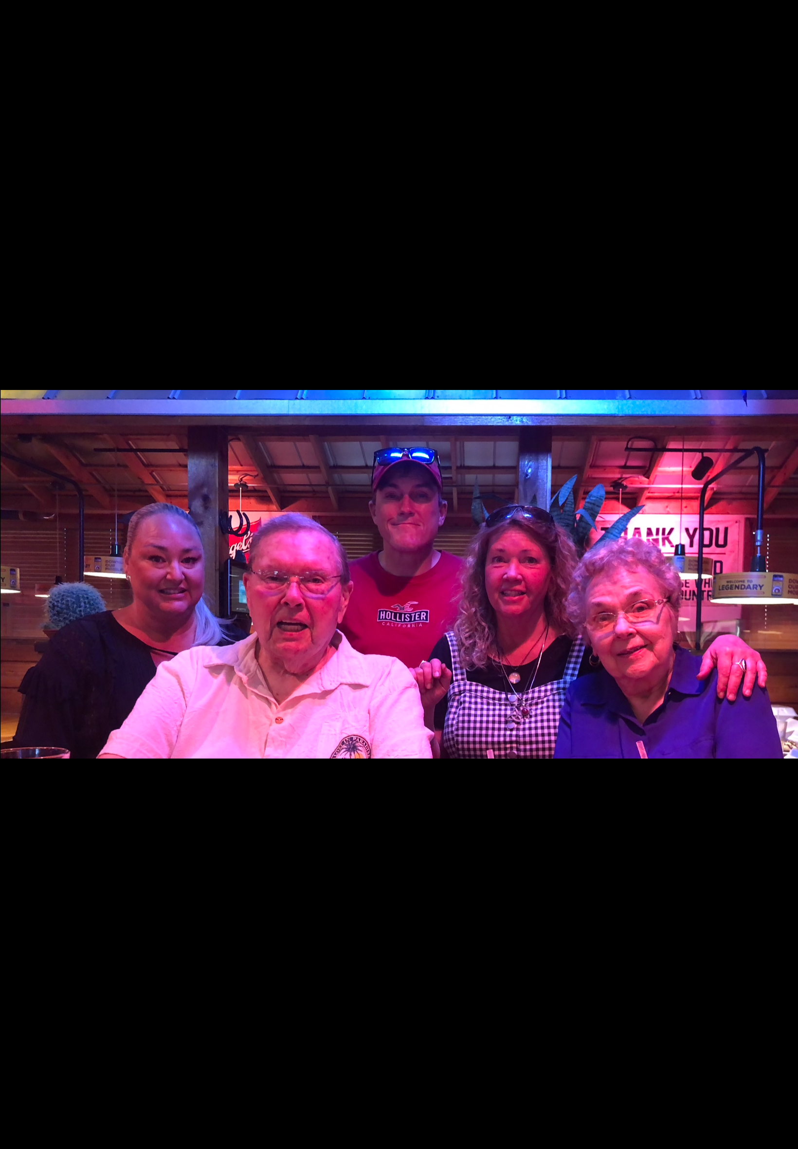 Six adults smiling for a group photo at a casual bar or restaurant with blue and pink neon lighting and wood decor behind them.