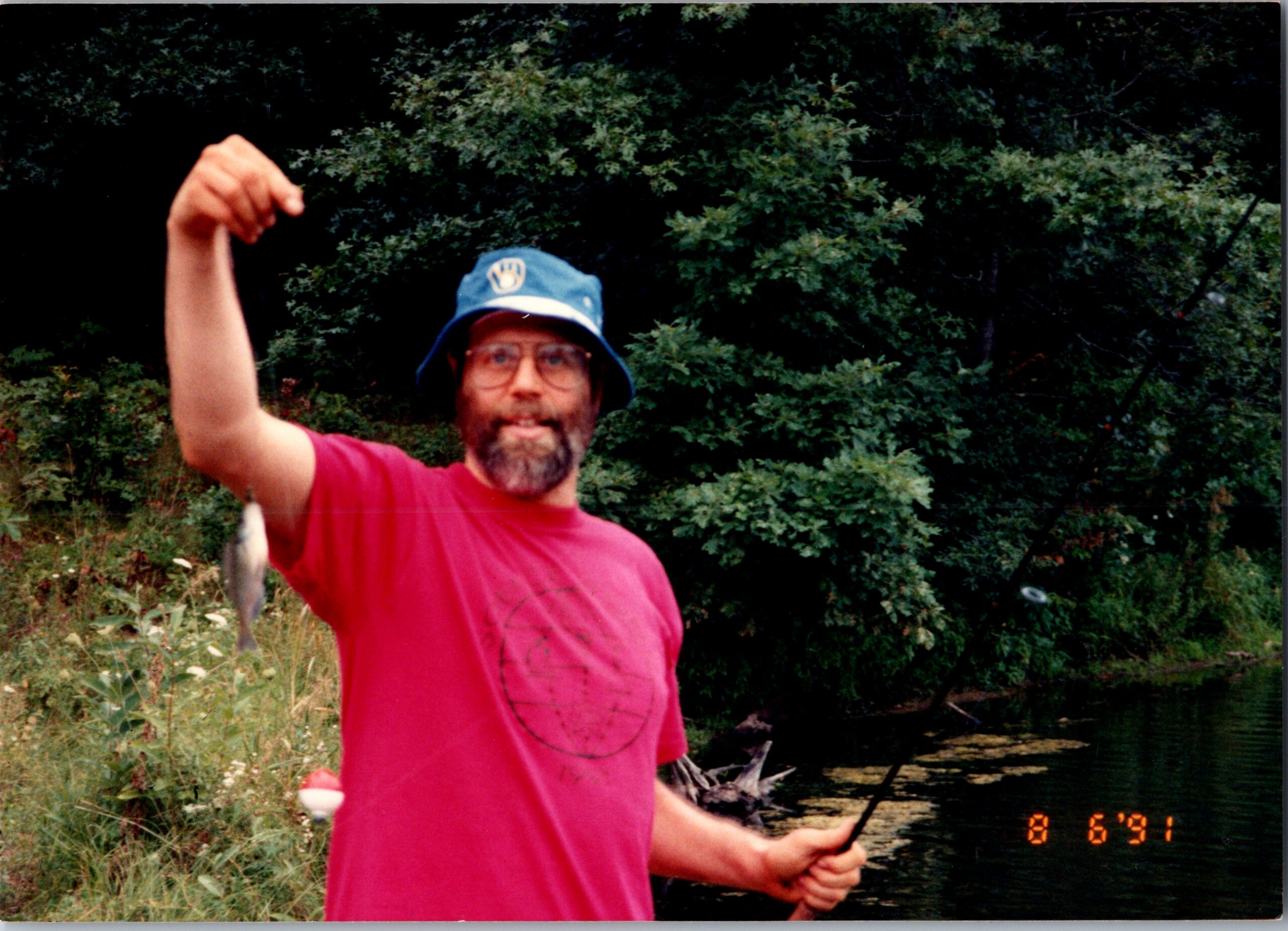 Man with a blue bucket hat and red shirt raises his fist outdoors by a wooded riverbank.